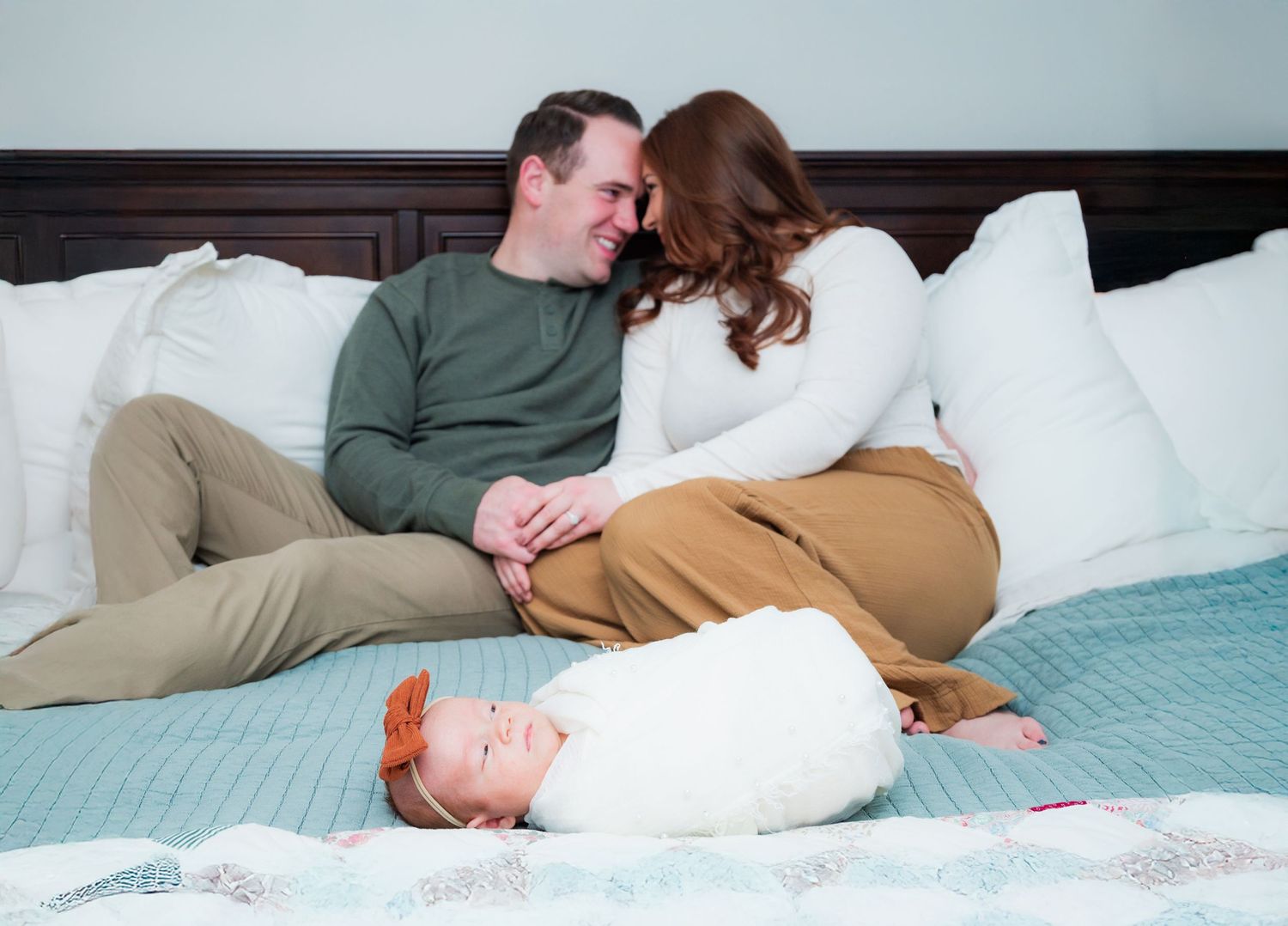 Family relaxes on bed with newborn baby in white swaddle.