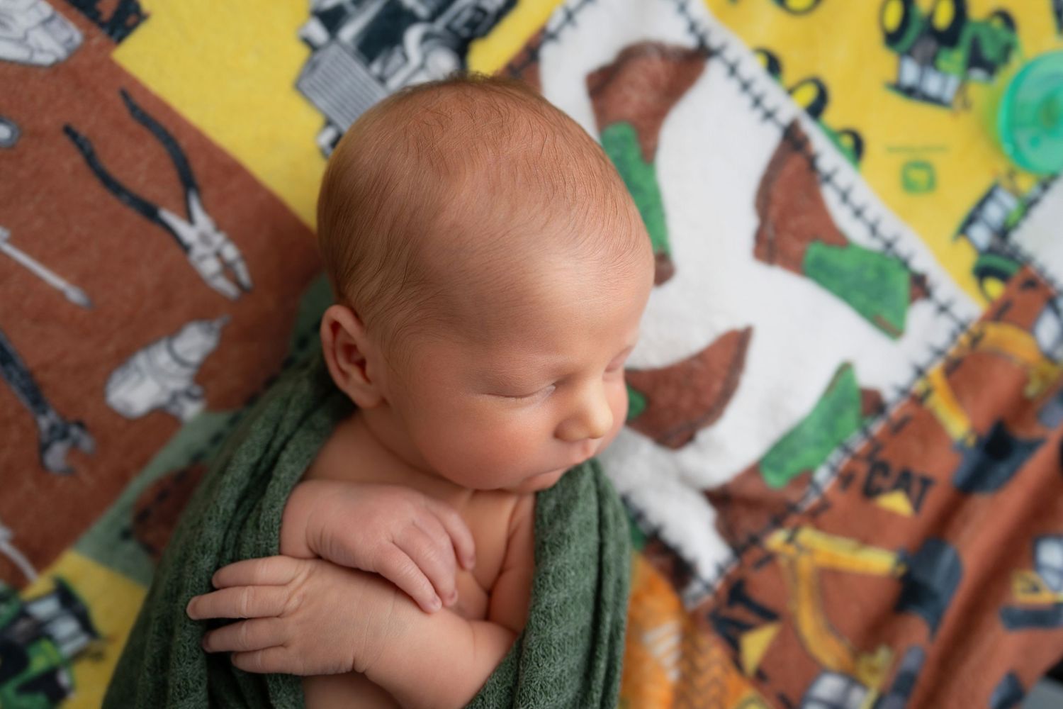 Newborn baby sleeping peacefully wrapped in green swaddle on colorful tools patterned quilt blanket.