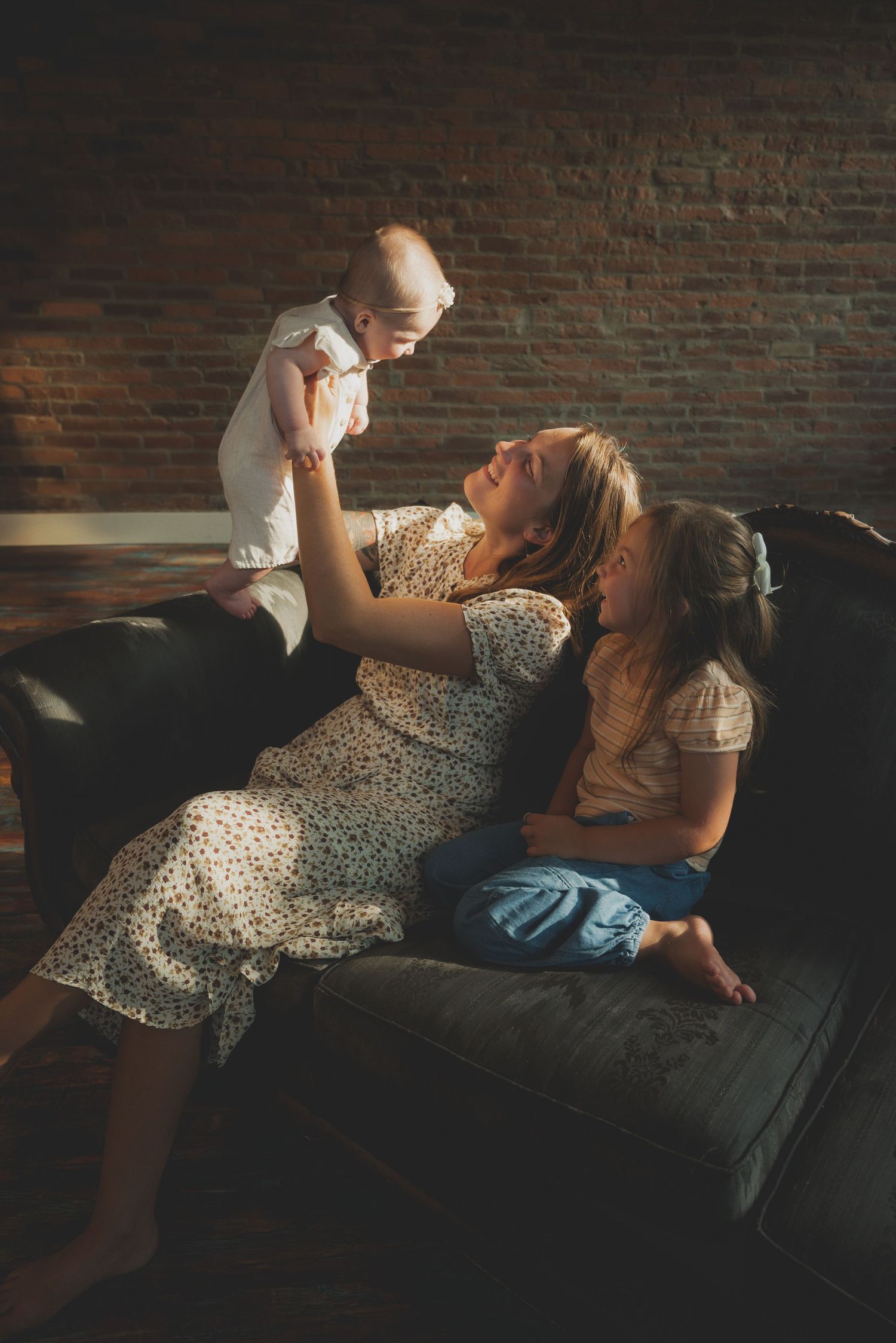 Parent and baby sharing playful moments on a dark leather couch in natural lighting.