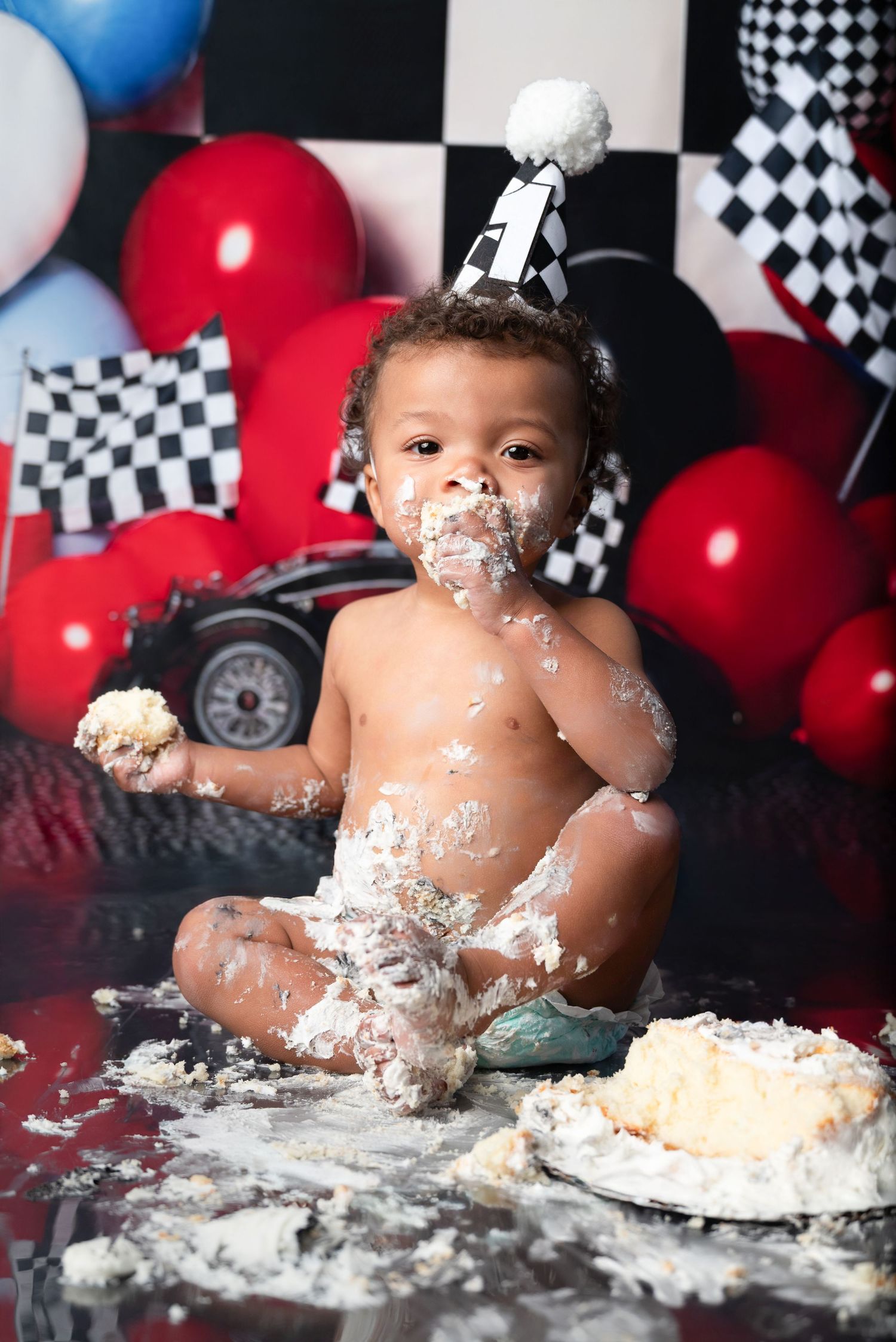Cake smash photo session with racing theme featuring balloons and checkered backdrop.