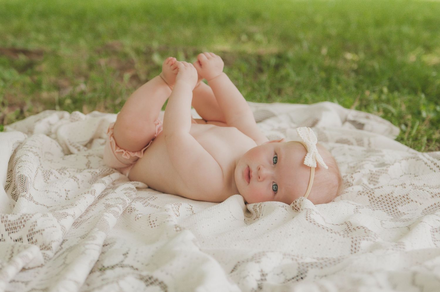 Baby lying on white lace blanket in grass during outdoor photo session.