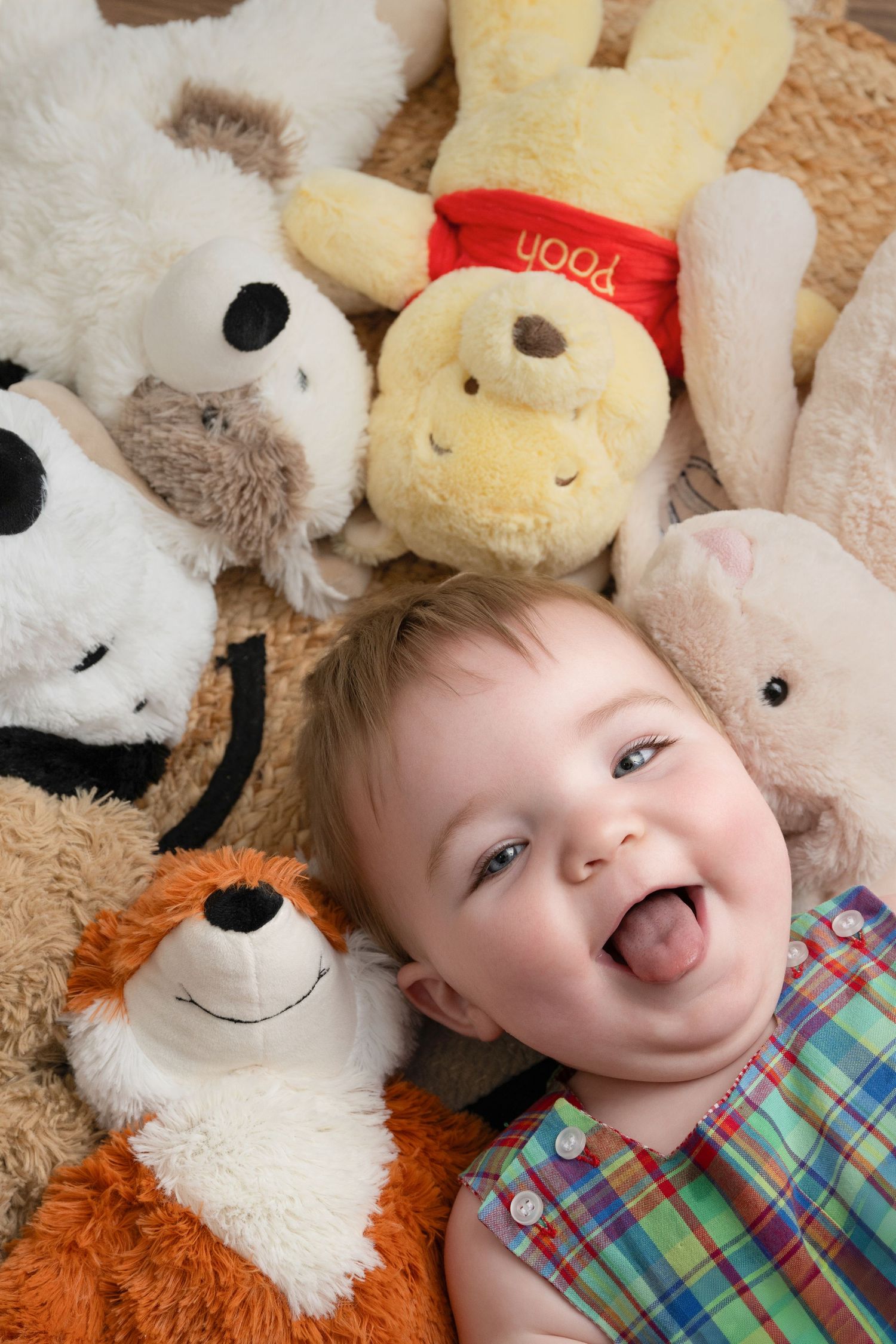 Happy baby laughing among stuffed animals including Winnie the Pooh and Snoopy plush toys on a soft carpet.