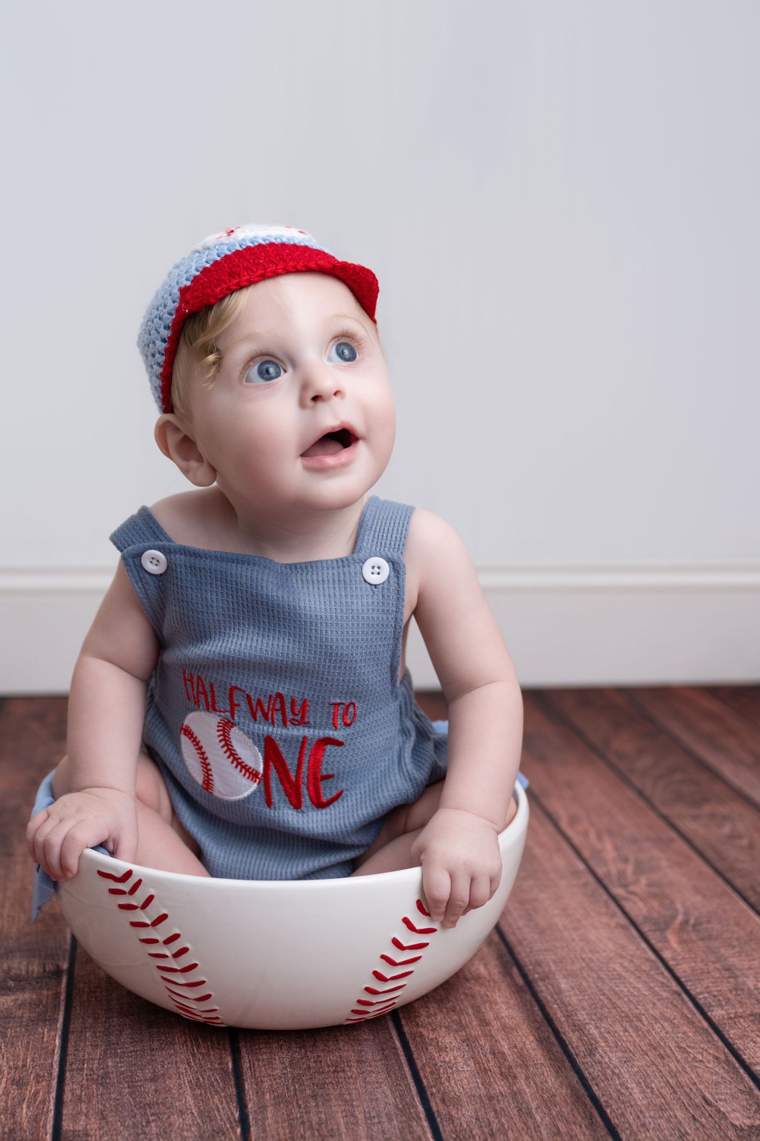 Adorable baby sitting in oversized baseball bowl wearing red cap and halfway to one romper on wood floor.