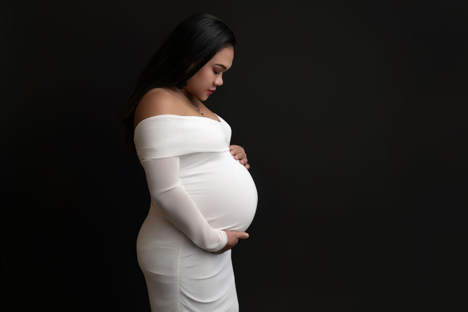 A maternity photo featuring an elegant white off-shoulder dress against a dark background in dramatic lighting.