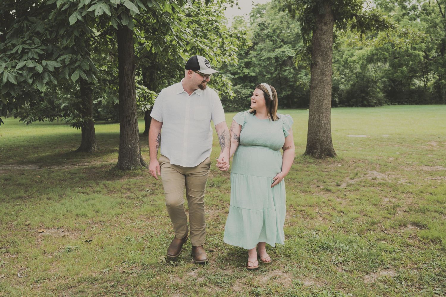 A couple walks hand-in-hand through a lush green park, the woman in a mint dress and man in a gray shirt.