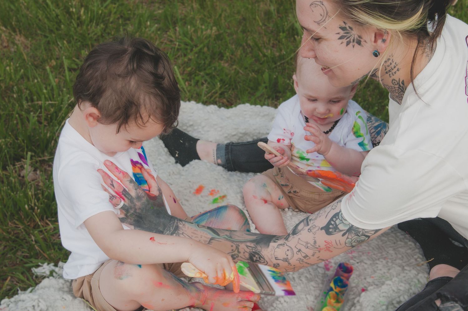 Mom and her babies sit together on grass while doing a colorful painting activity outdoors.