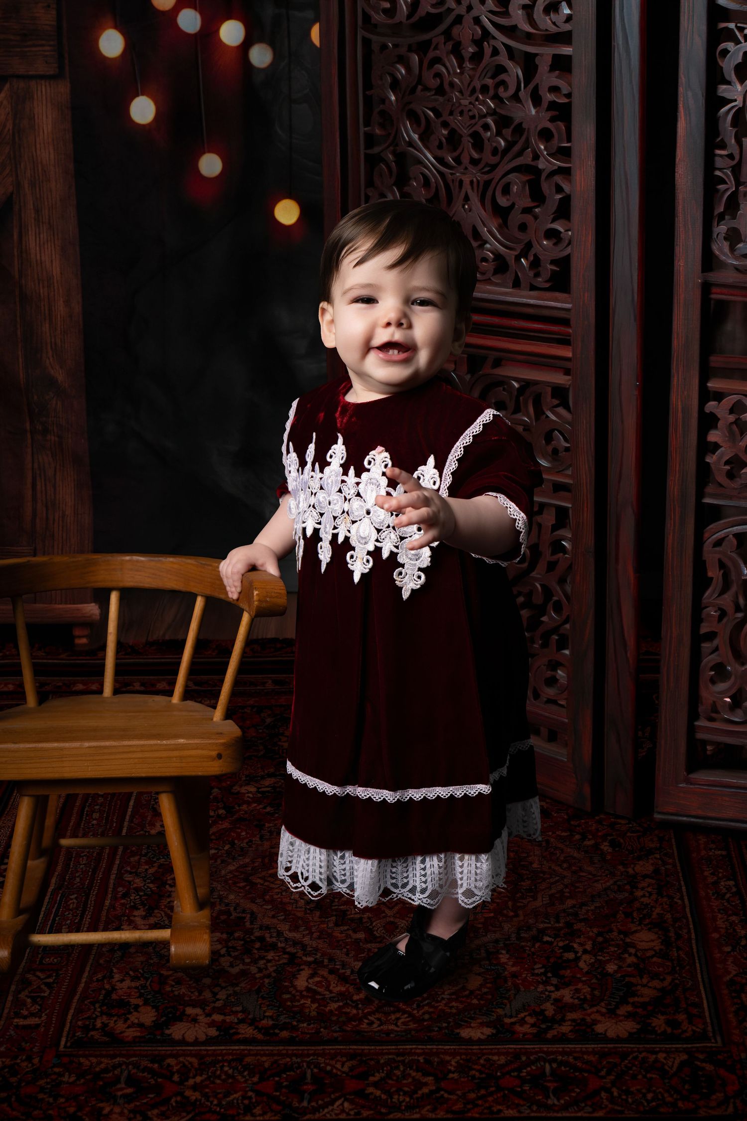A small child in a black dress with white lace trim sits in a dark wooden chair against an ornate background.