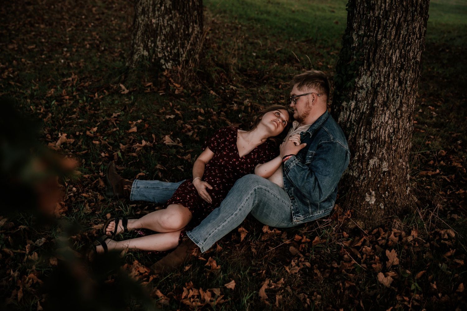 Couple sitting together on fallen autumn leaves beneath trees. Maternity fall photos SOKY Franklin KY