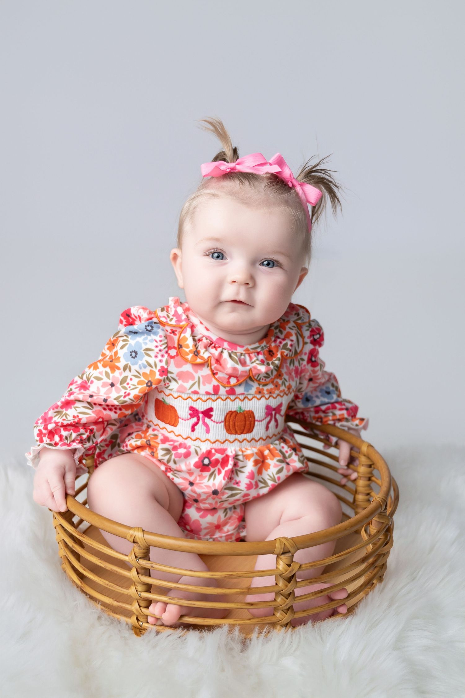 Baby wearing floral dress sits in woven basket during professional photo session.