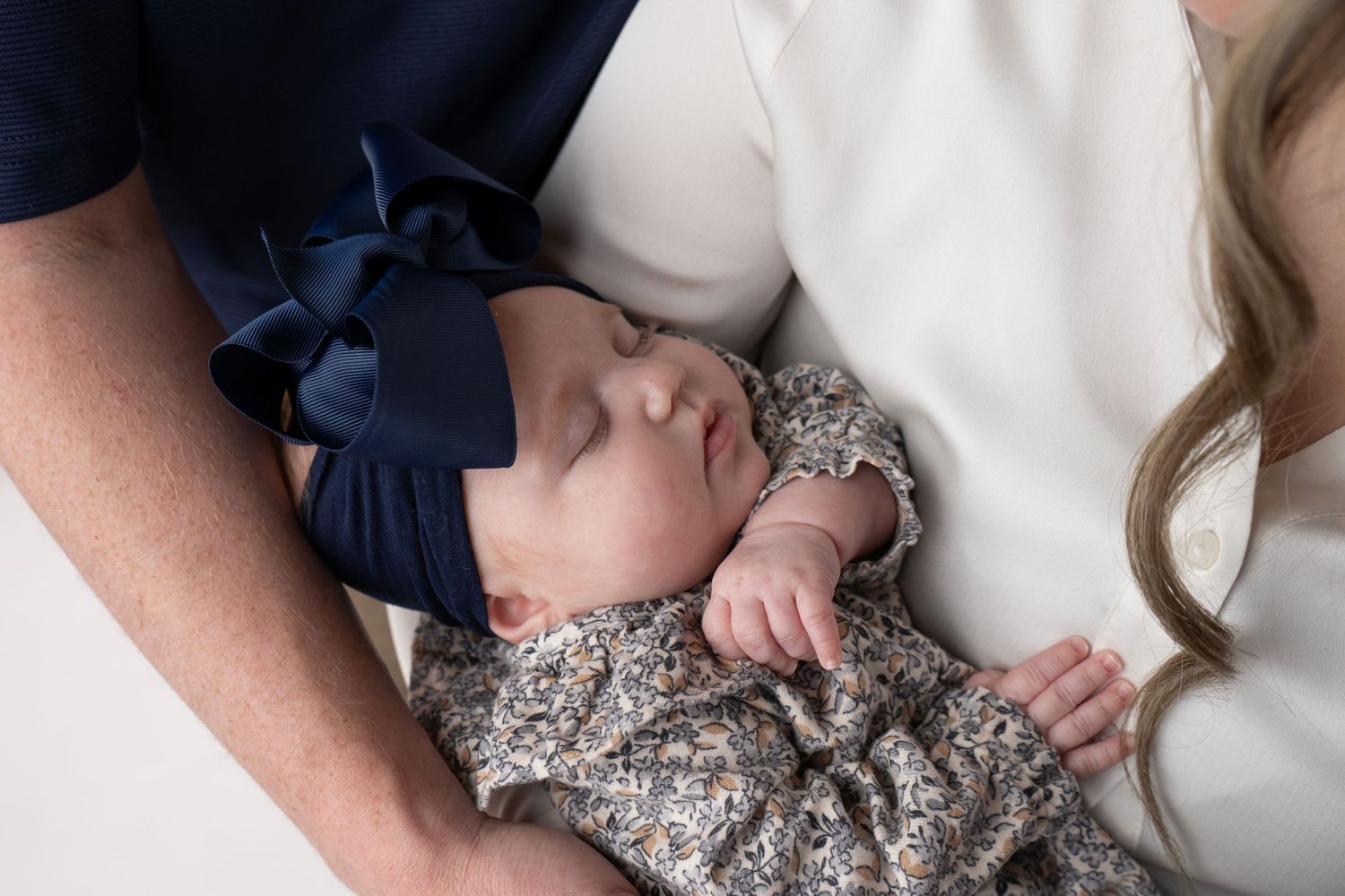 A mother in a navy dress holds and comforts her baby wearing a floral print outfit against a white backdrop.