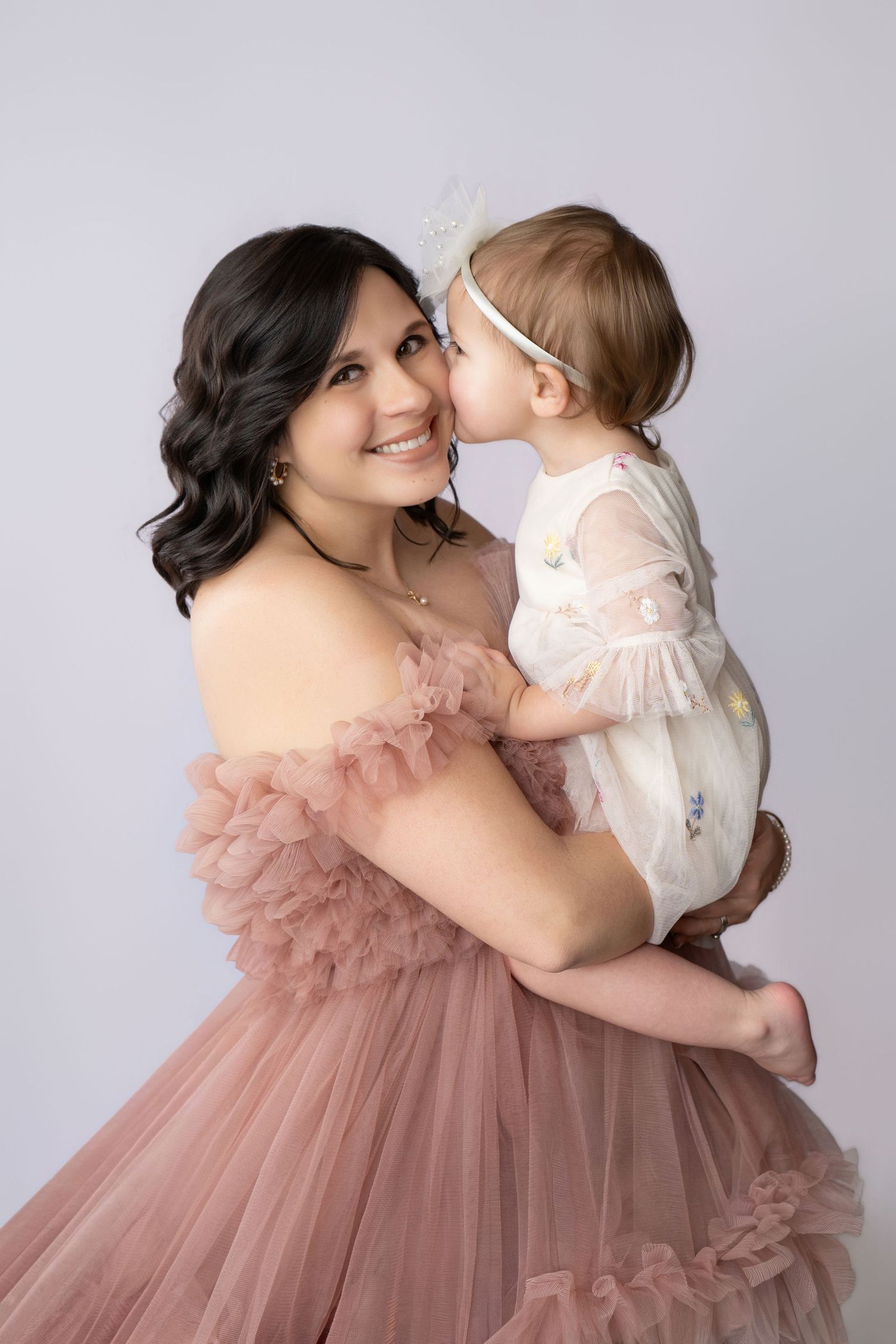 A smiling woman in a pink tulle gown holds a baby in a white floral romper who kisses her cheek.