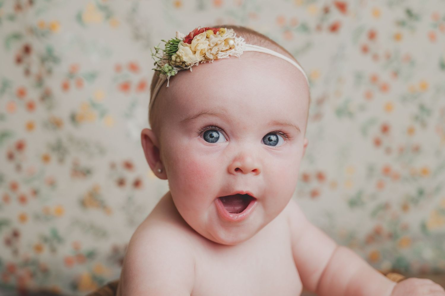 Baby portrait with floral backdrop and delicate headpiece in soft lighting.