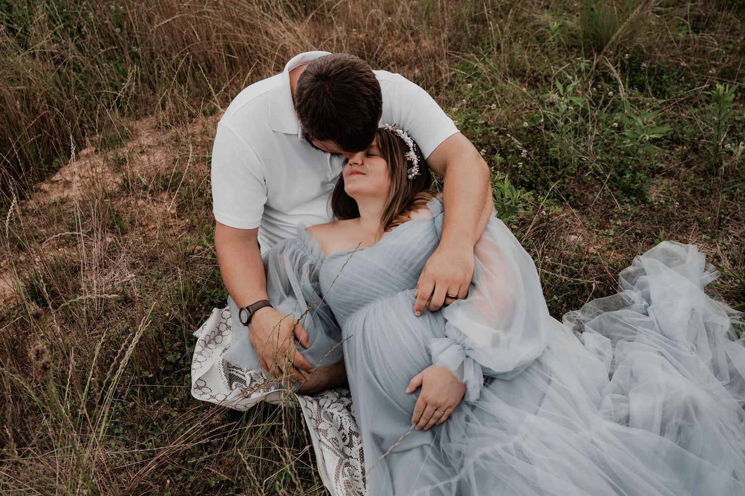 A couple shares a tender moment in a field, the woman in a flowing blue gown resting as her partner leans in to kiss her.