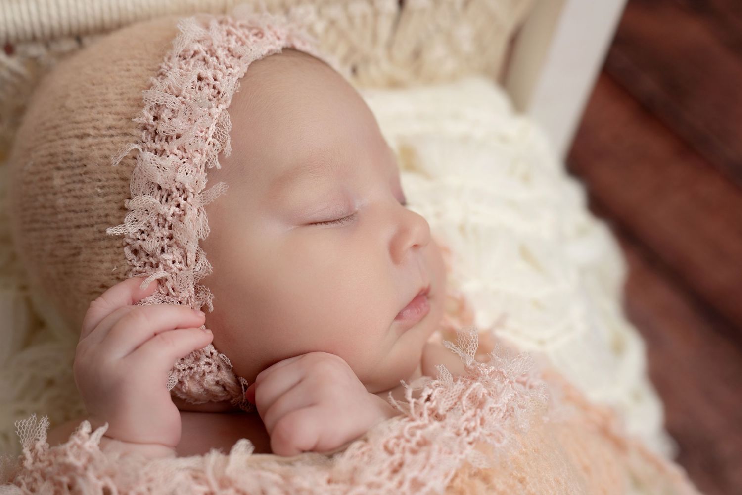 Sleeping newborn wearing delicate pink lace bonnet rests peacefully on soft white blanket during professional photo session.