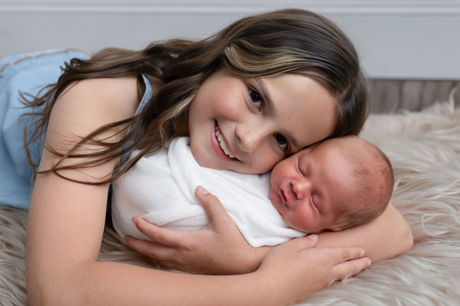 Smiling young girl lying down and gently holding a swaddled newborn baby on a fluffy white rug.