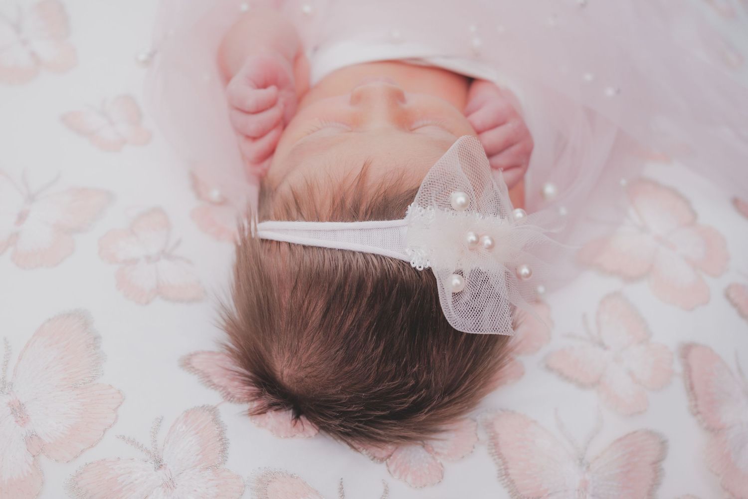 Newborn baby wearing a delicate white bow headband lying on pink butterfly bedding.