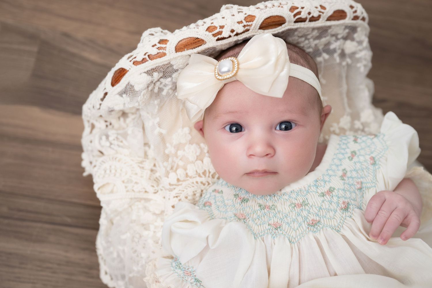 A newborn baby in a cream lace gown and bow headband rests in a decorative white wicker bassinet.