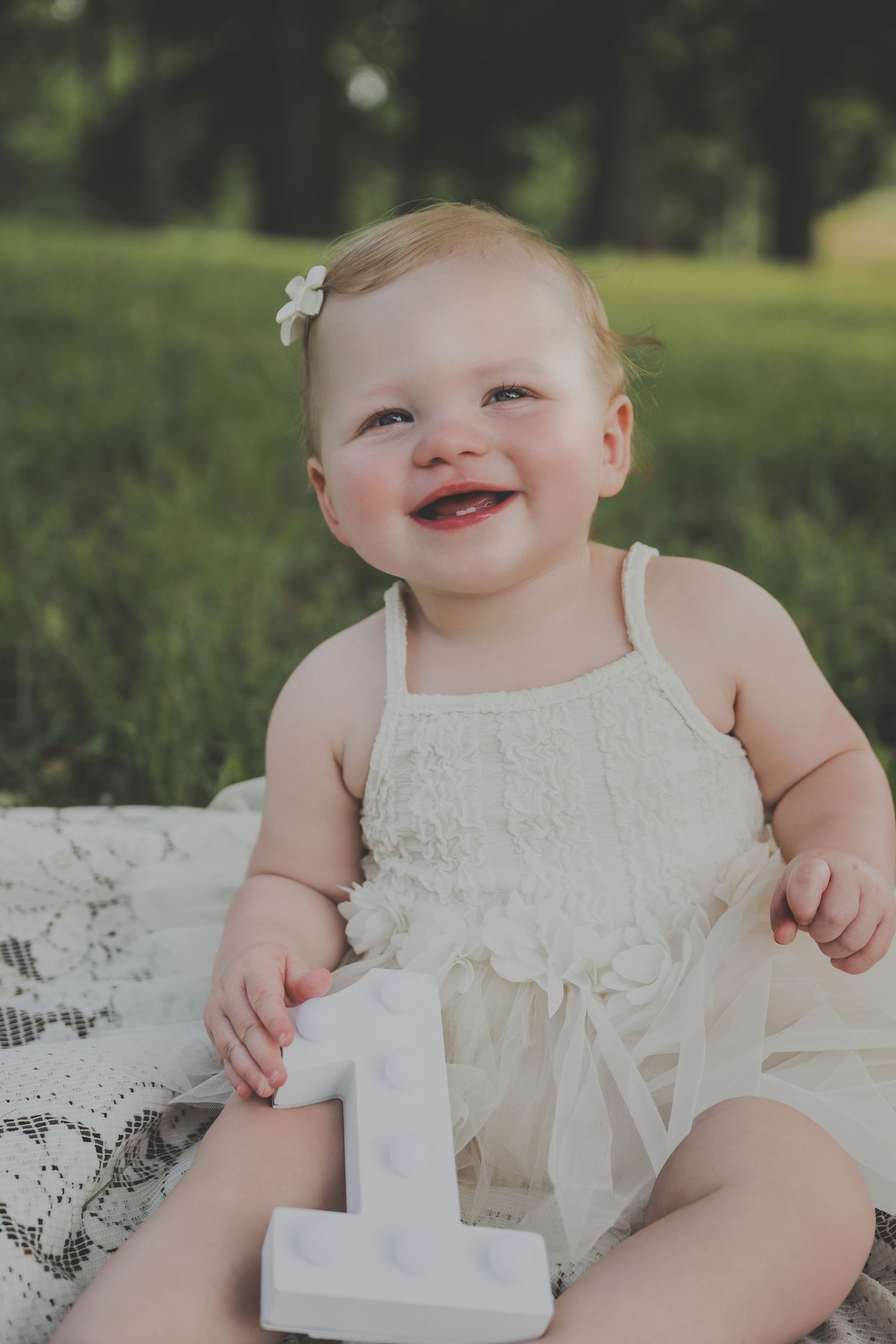 Smiling baby girl in white dress sitting on lace blanket outdoors, celebrating her first birthday with number one block.