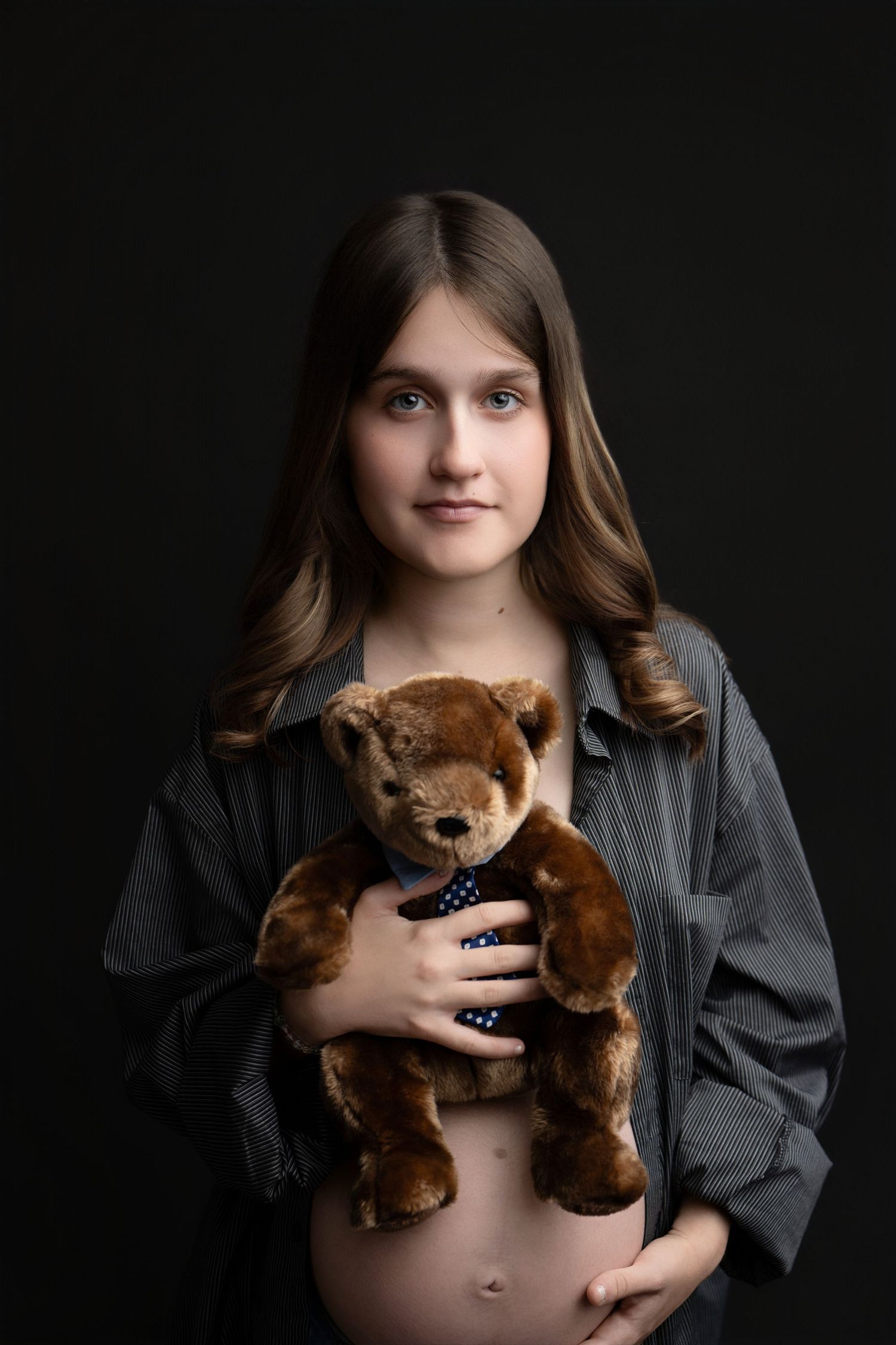 Artistic maternity portrait with woman holding brown teddy bear against dark backdrop.