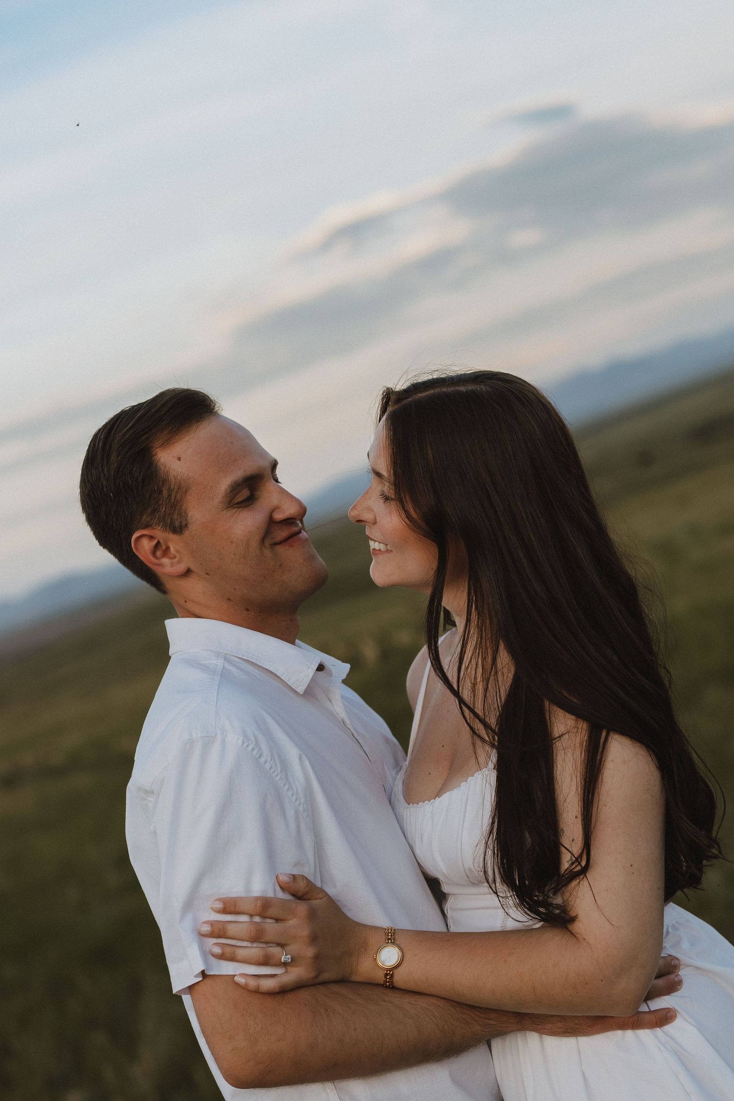 A couple in white outfits embracing and smiling at each other against a scenic mountain landscape at dusk.