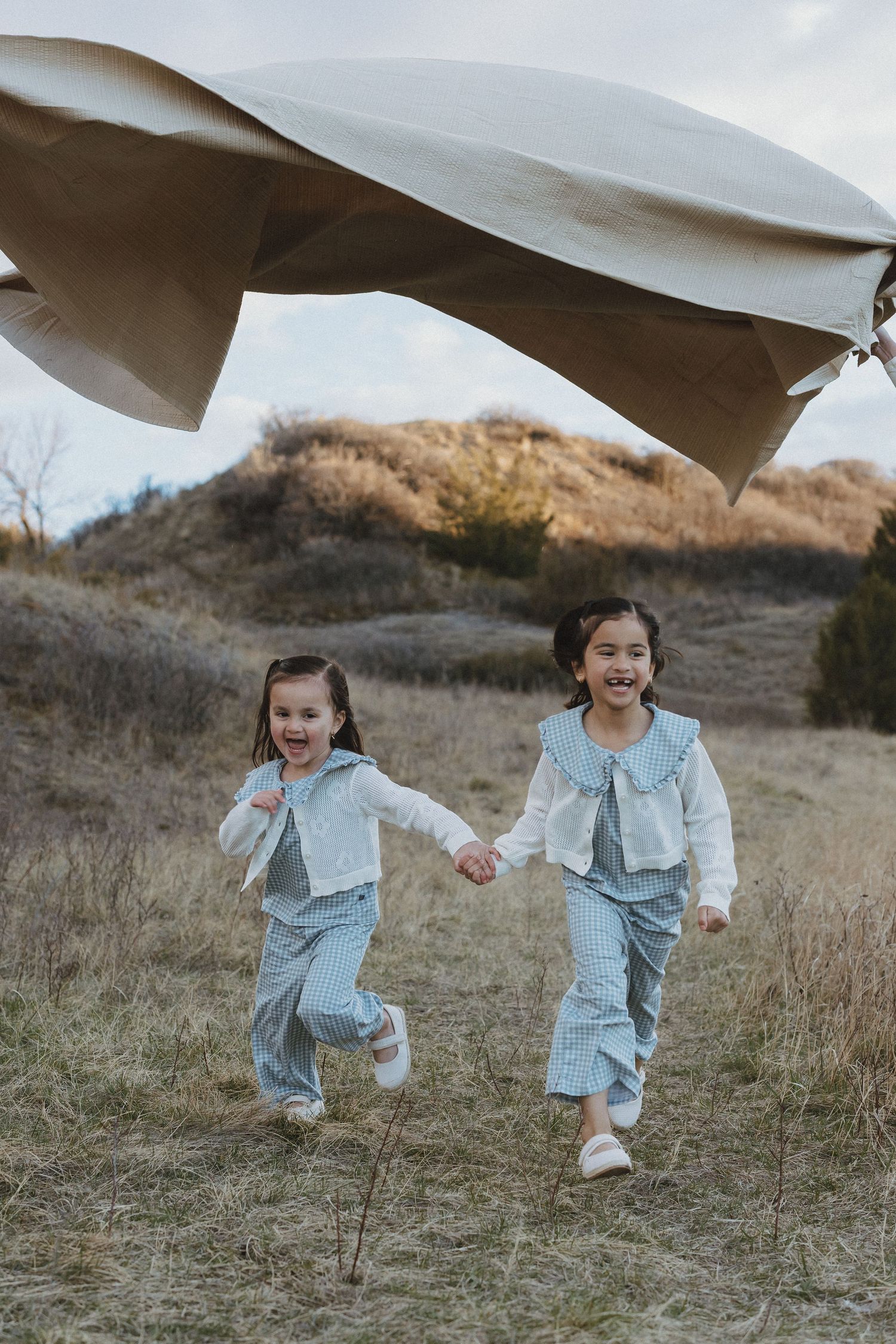 Two young girls in matching blue outfits hold hands running through a field with a fabric piece flying above them.