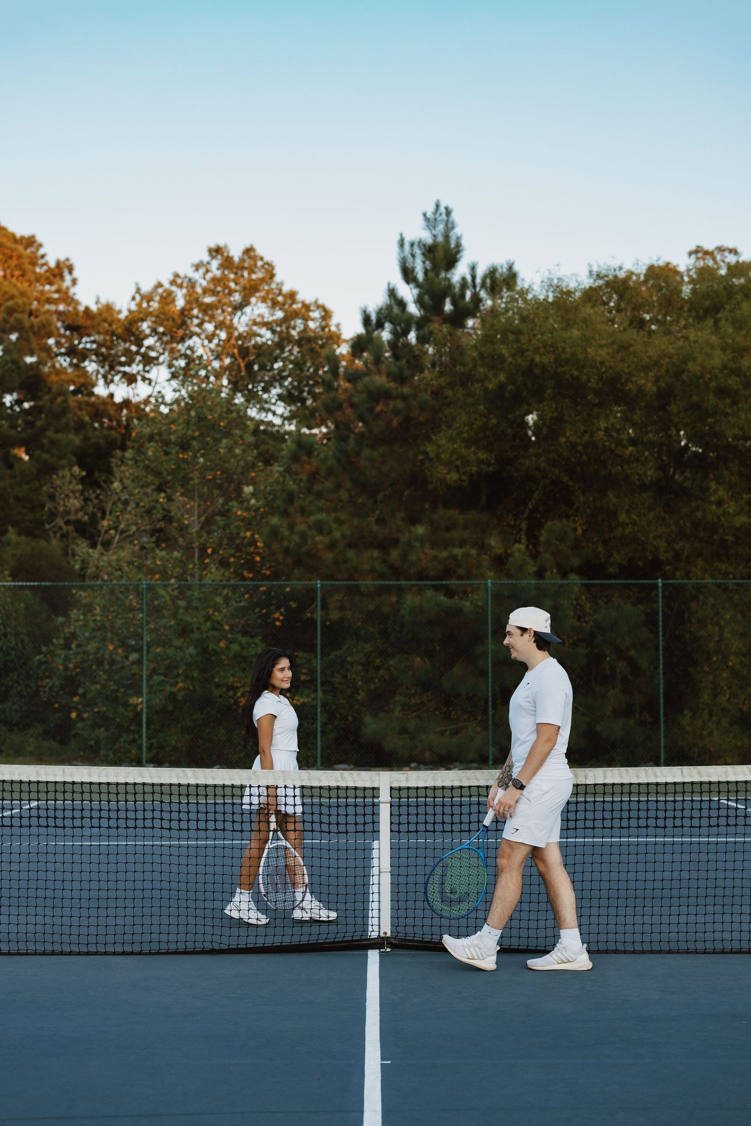 People playing tennis on an outdoor blue court at sunset with trees in the background.