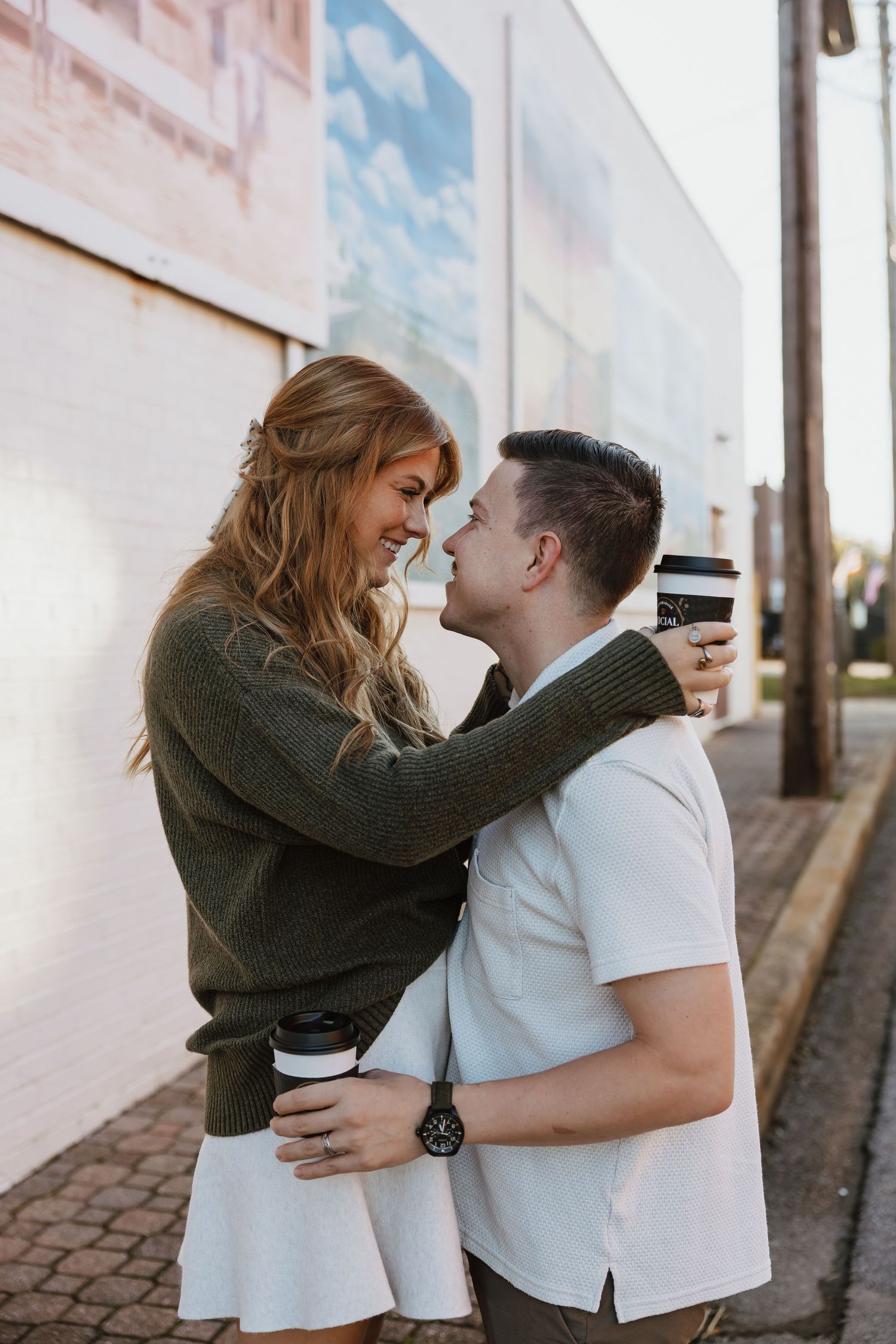 A couple in white and olive green outfits share an intimate moment against a white wall on cobblestone street.