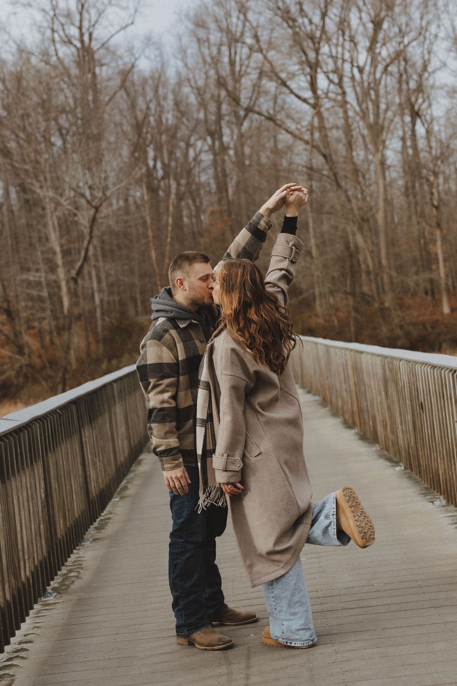 Couple dancing together on a wooden bridge surrounded by bare autumn trees in a romantic outdoor setting.