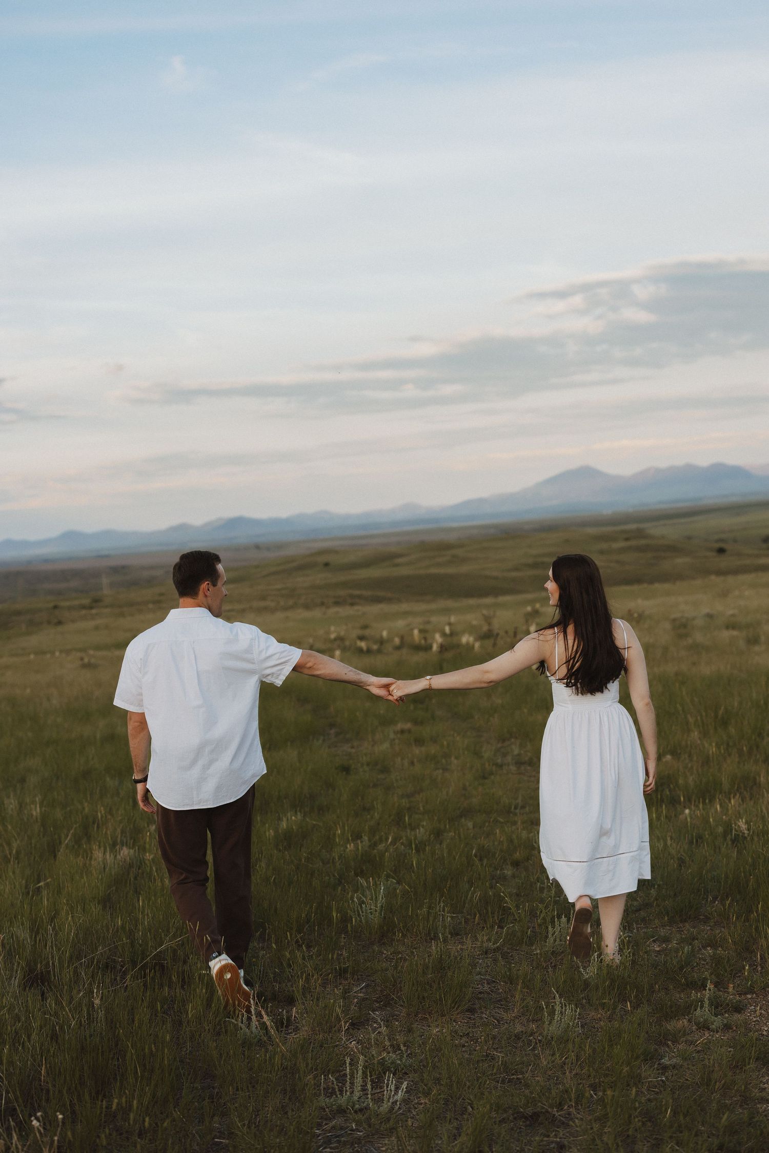 A couple holds hands walking through a lush green meadow with mountain views under a cloudy sky.