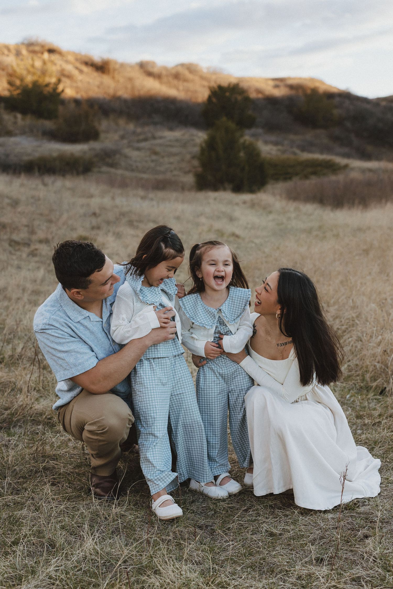 Family of four laughing together in a golden field at sunset, two girls in blue matching outfits with parents in casual attire.