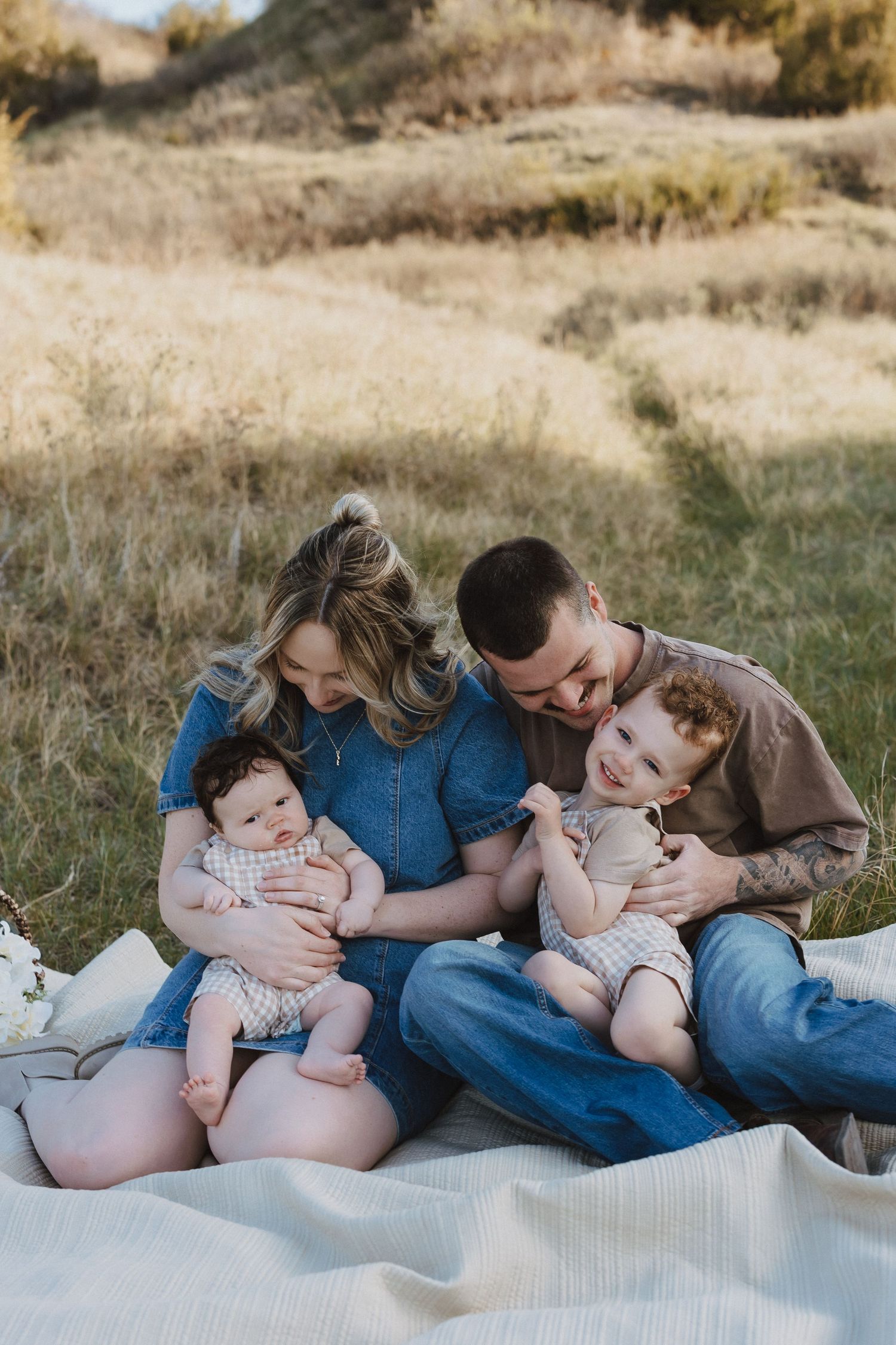 Family of four sitting on blanket in golden field, parents holding two babies in matching outfits during outdoor photo session.