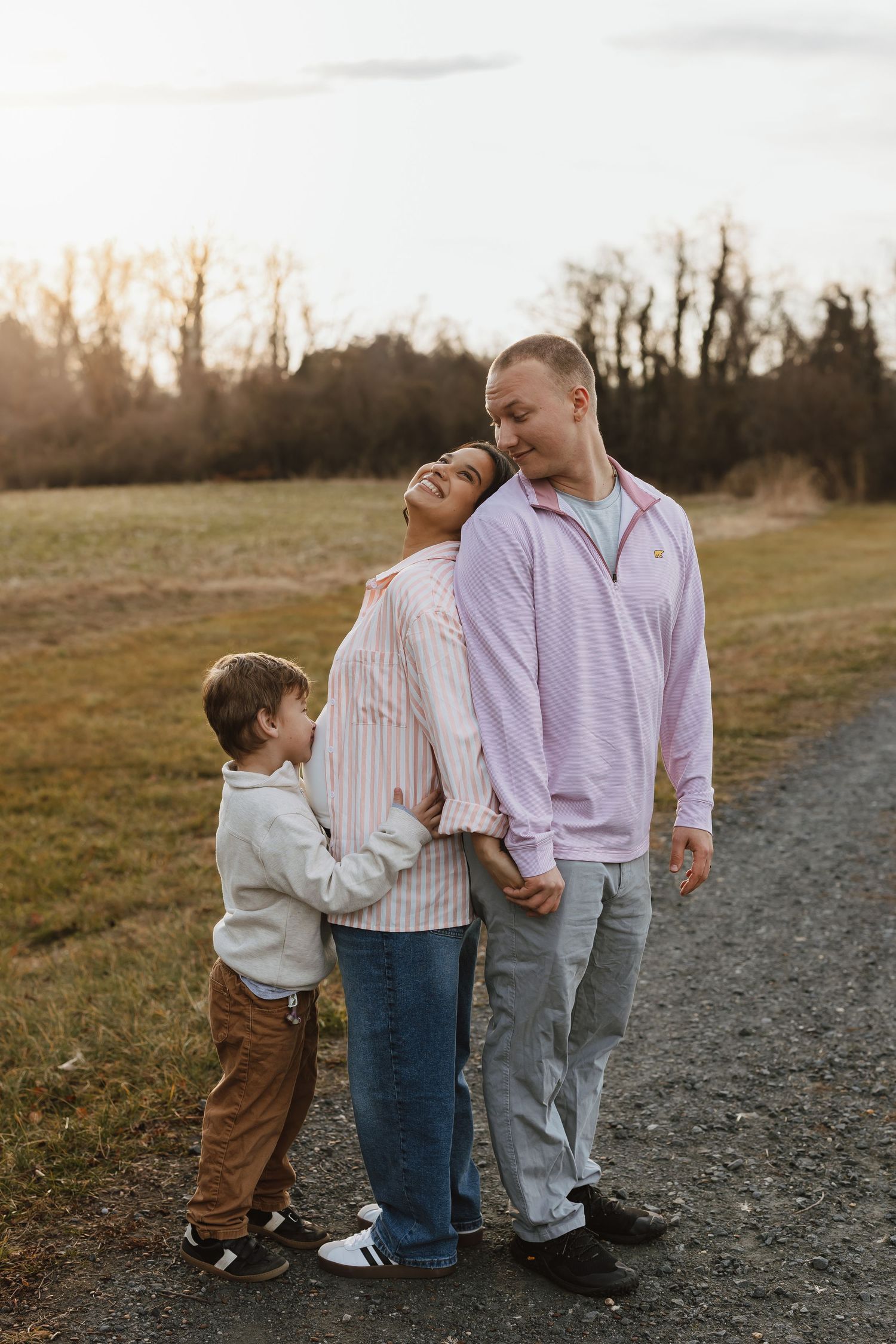 A family of three stands together on a gravel path at sunset while their young child hugs them from behind.