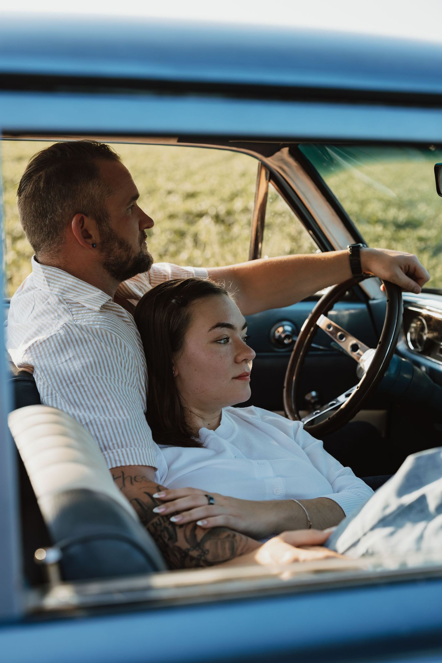 Two people sit in a vintage car with blue exterior, with one resting while the other is at the steering wheel.