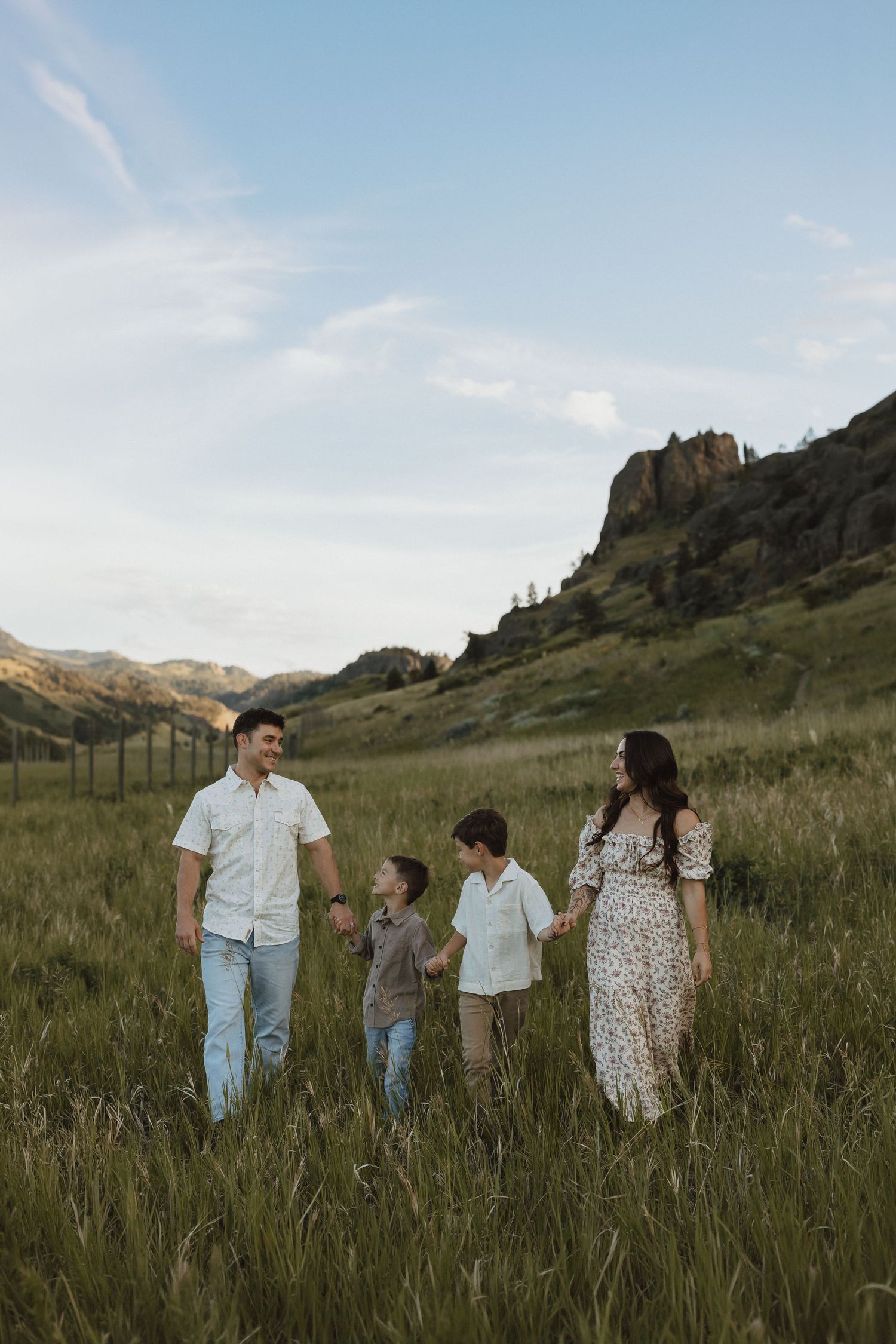 Family of four holding hands walking through a green meadow with rocky hills and blue sky in background.
