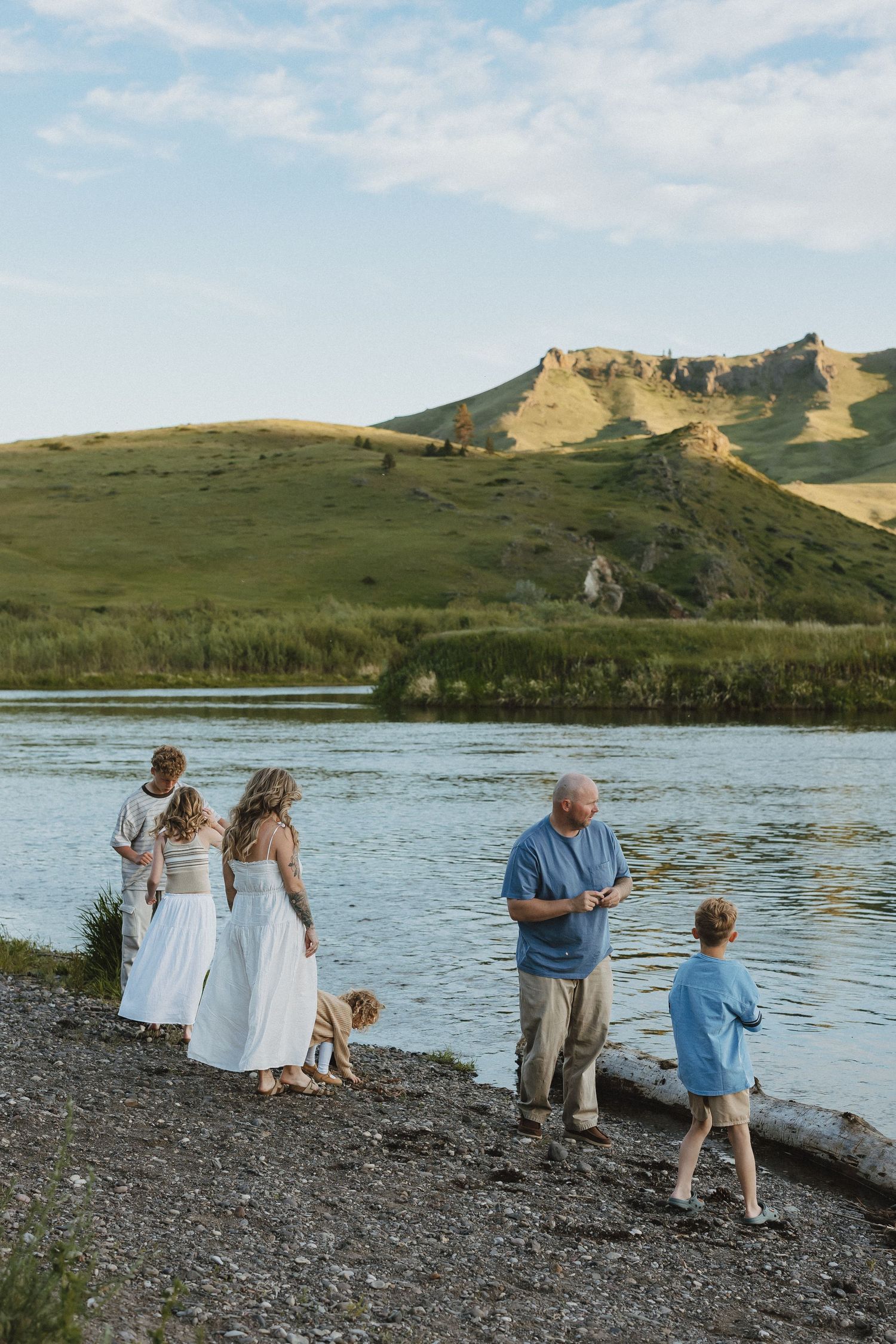 Family gathering by a scenic river with green hills, enjoying a sunny day near the water's edge.
