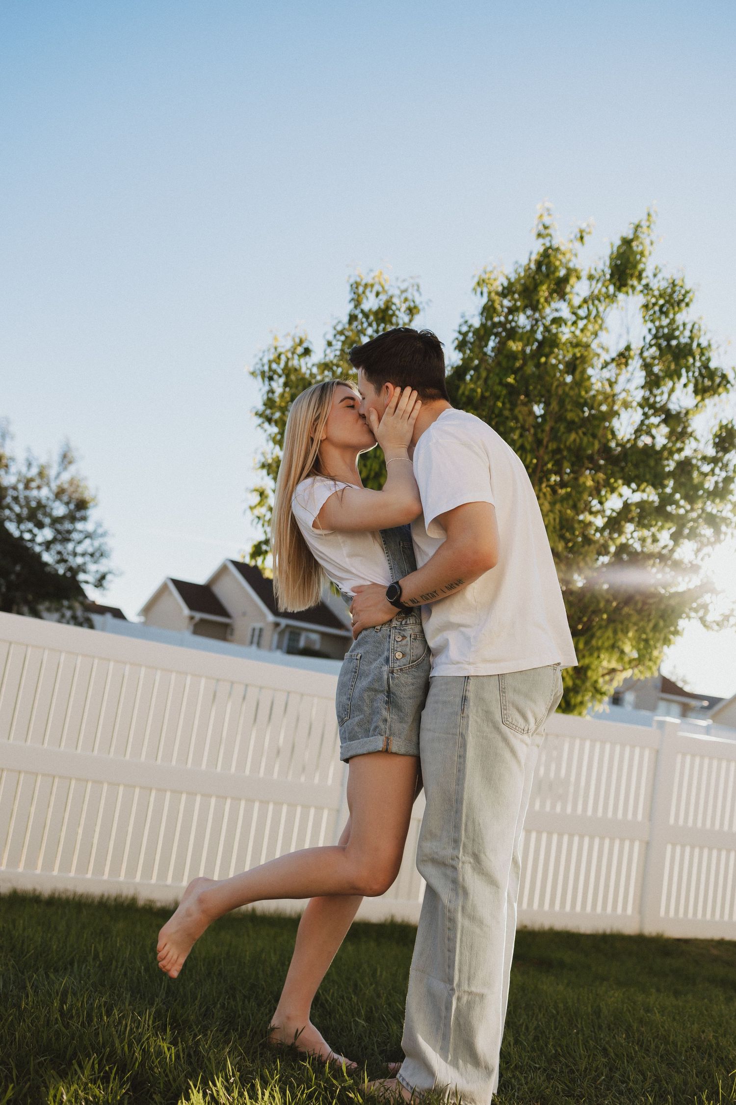 A couple sharing a romantic kiss in a sunny backyard with a white fence and green trees in the background.