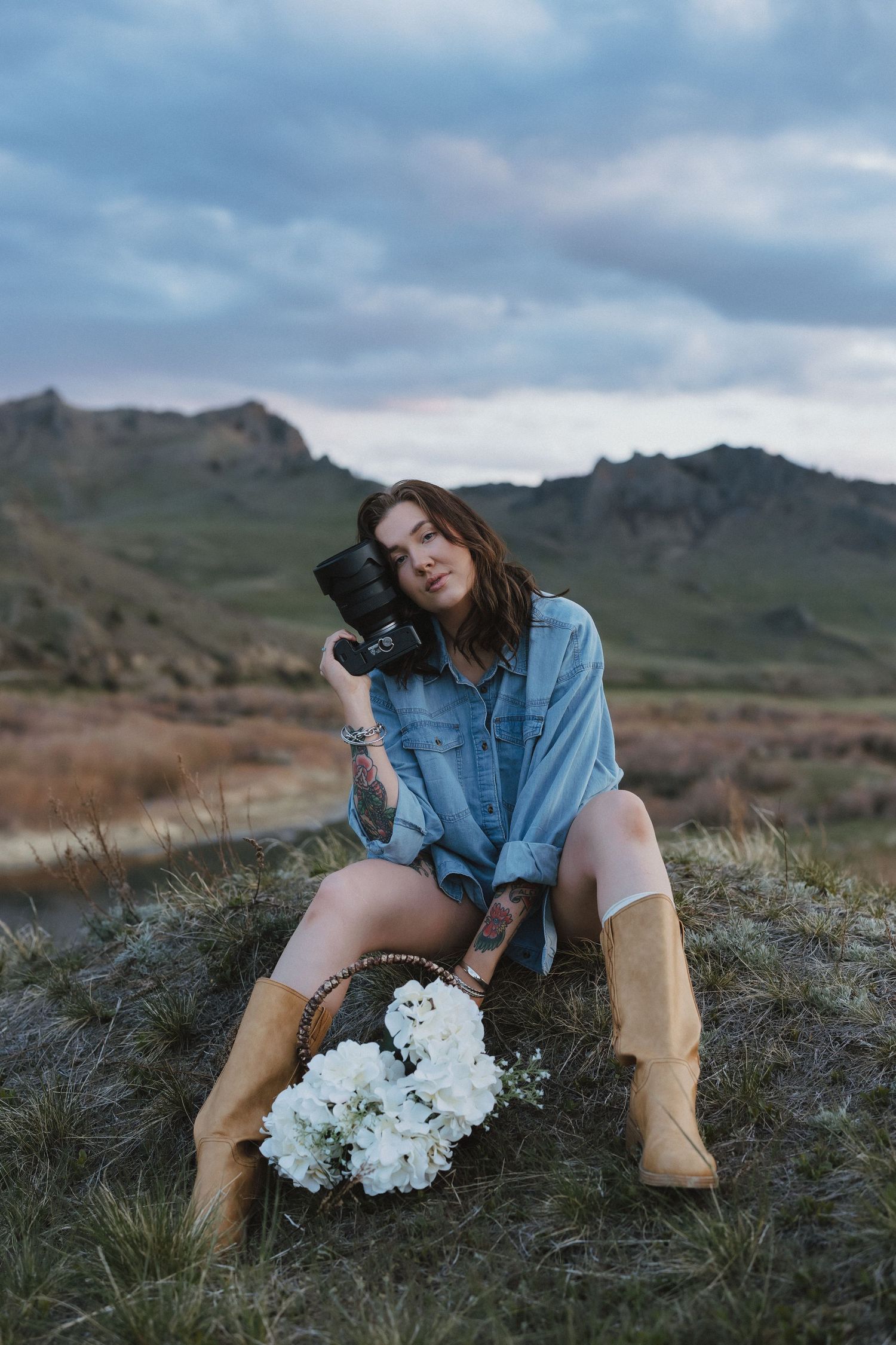 Woman in denim shirt and tan boots holds camera outdoors, with white flowers and scenic mountain landscape behind her.