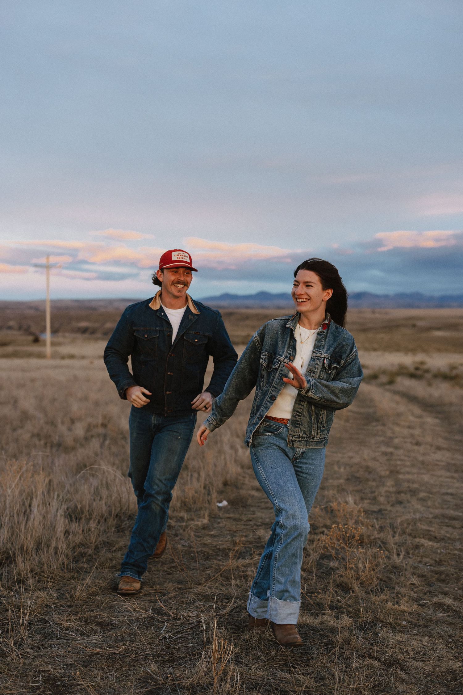 A couple walks together across an open field at sunset wearing casual denim outfits in a rural landscape.