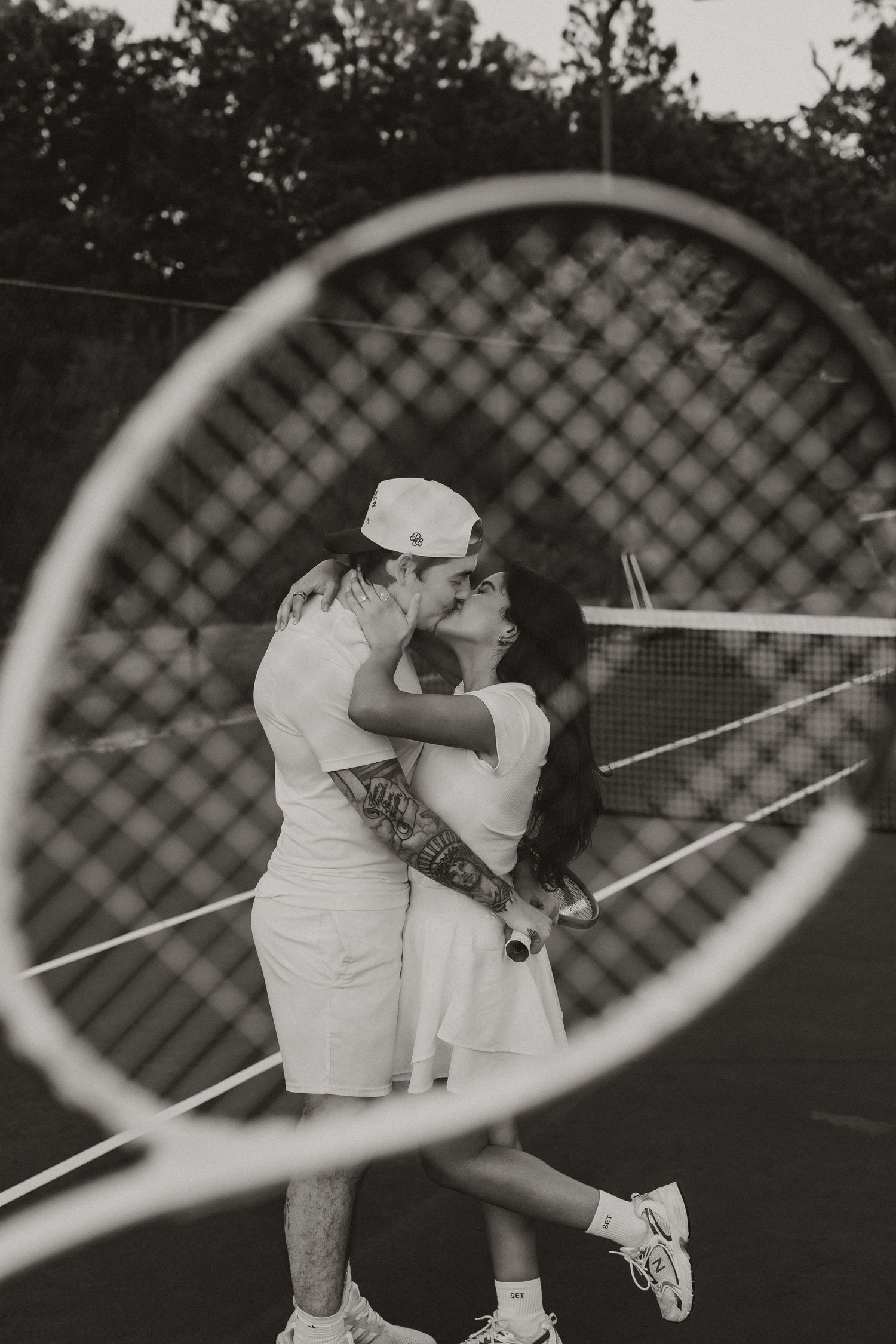 A romantic black and white photo of a couple embracing on a tennis court, framed through a tennis racket.