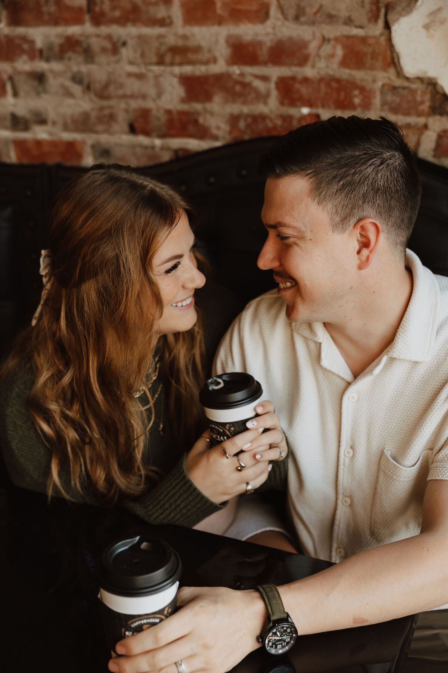 A couple sharing intimate moments and laughter while holding coffee cups against a rustic brick wall background.