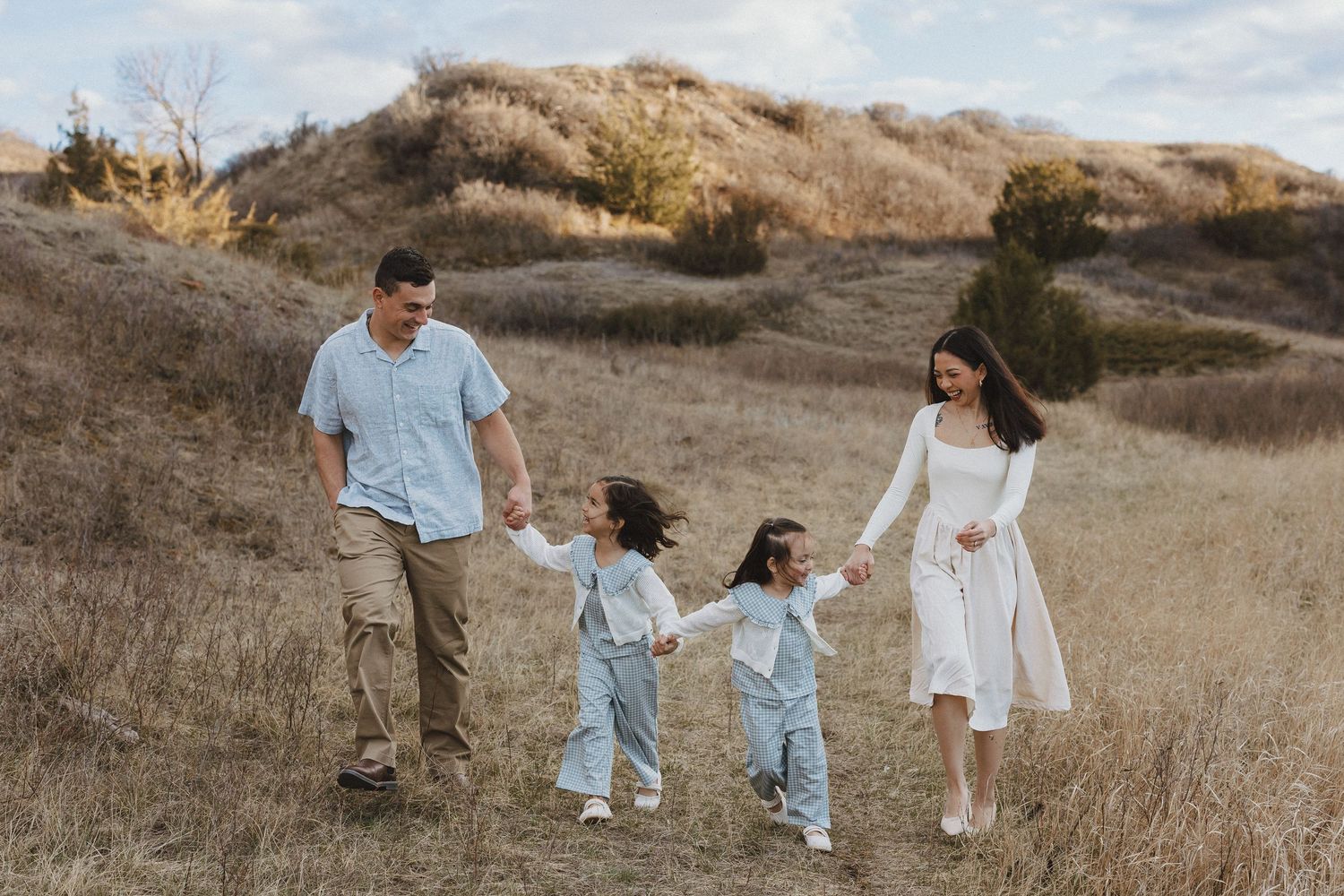 Family of four walking hand-in-hand through golden meadow, parents and two young children in light blue outfits.