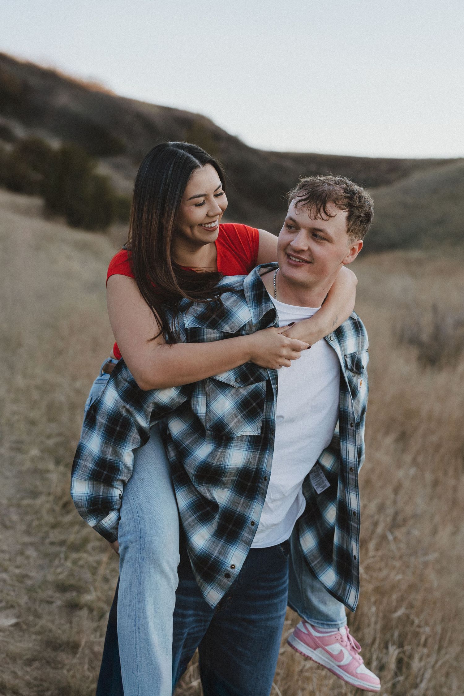 A joyful couple shares a piggyback ride in a scenic field, both smiling and wearing casual outfits.