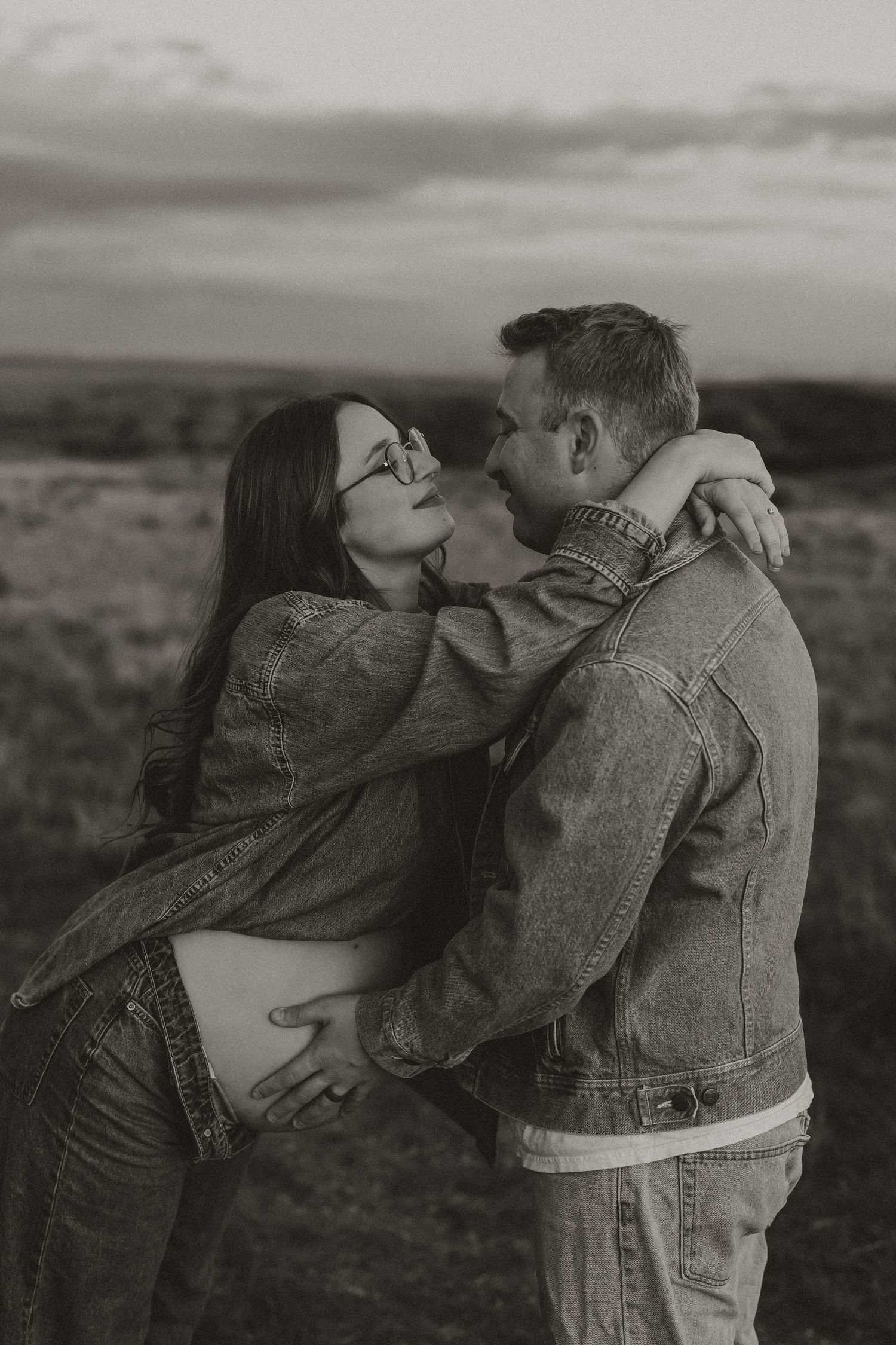 Black and white photo of a pregnant couple embracing outdoors at dusk, woman with glasses leaning into mans arms.