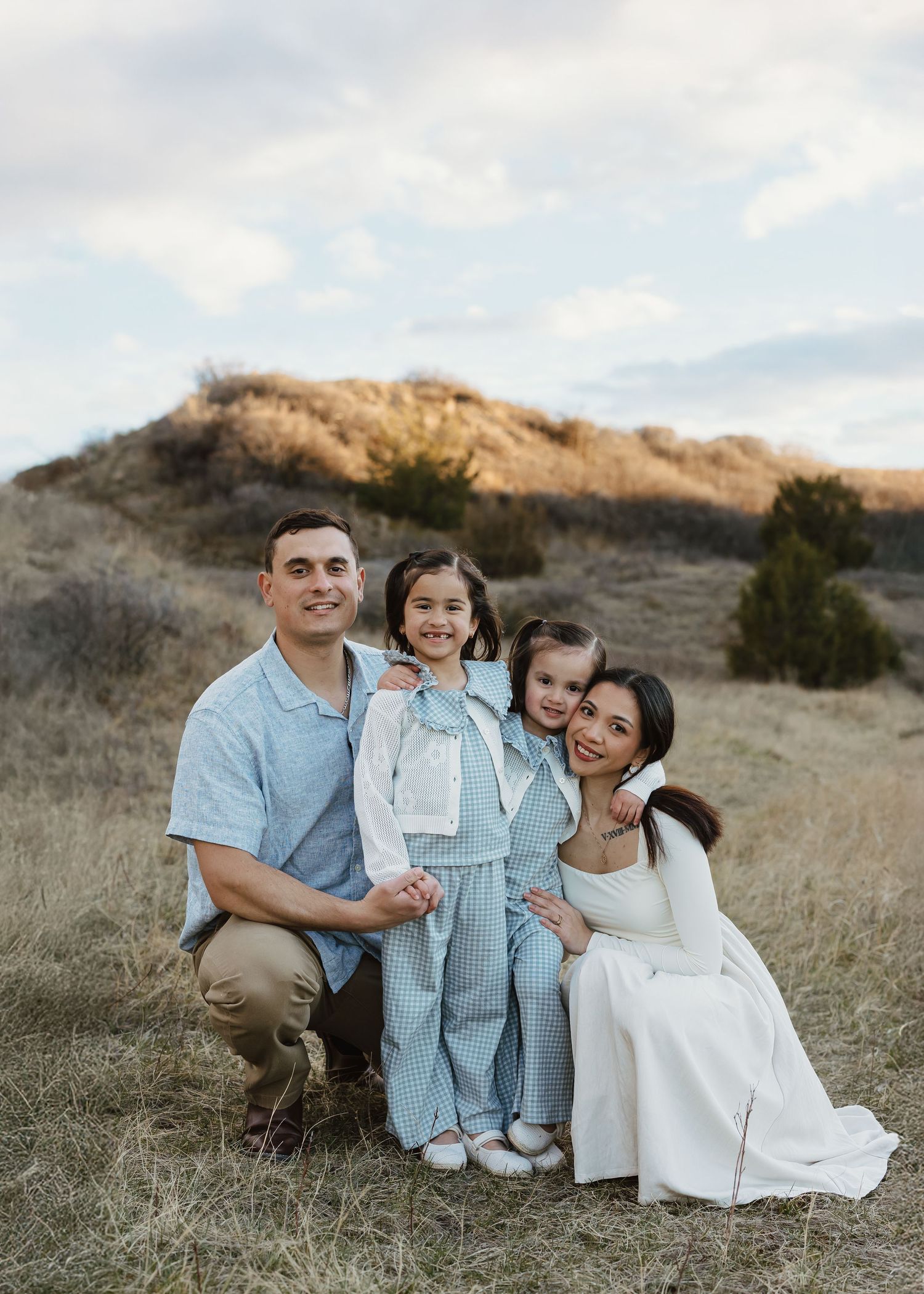 Family of four poses outdoors in a golden field, parents smiling with two young daughters in coordinating blue and white outfits.