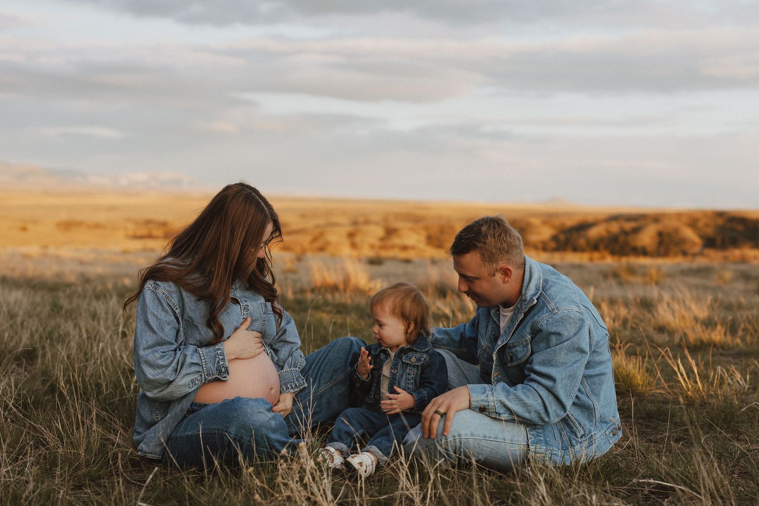 Pregnant woman and man sitting in golden field with toddler, family maternity photo session at sunset.