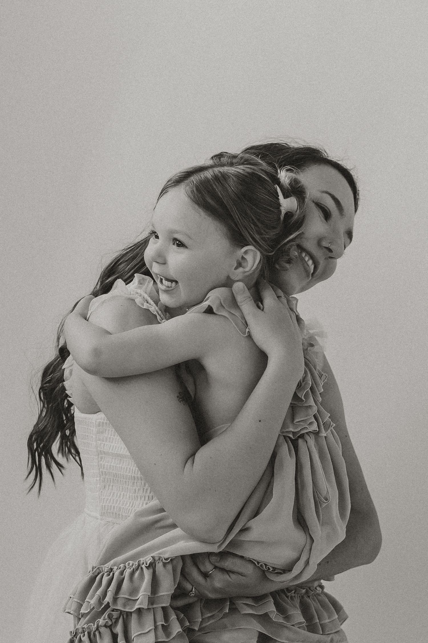 Black and white photo of a woman and young girl hugging and laughing joyfully in matching ruffled dresses.