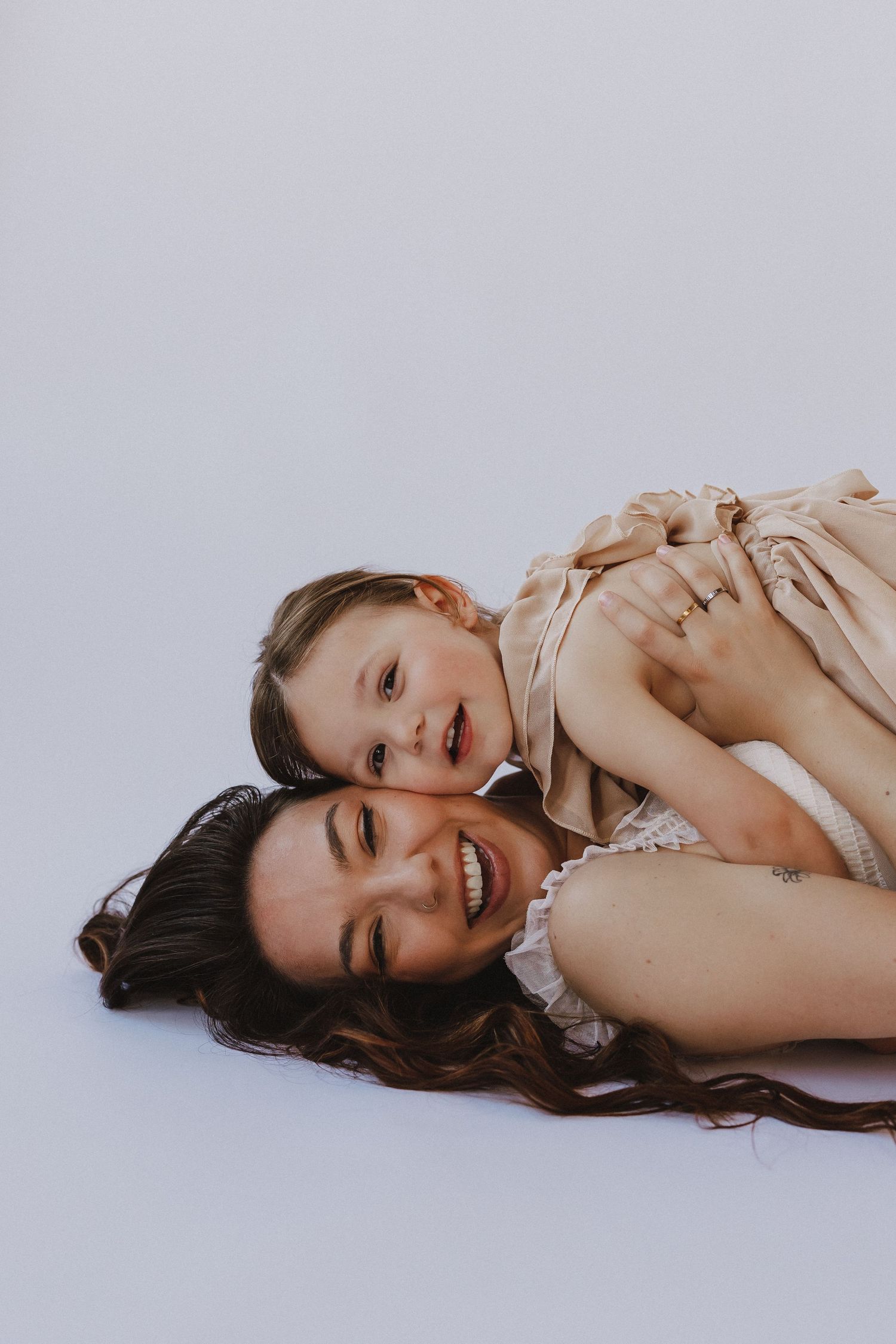 A smiling mother and young daughter lying together on a white background, wearing matching beige outfits.
