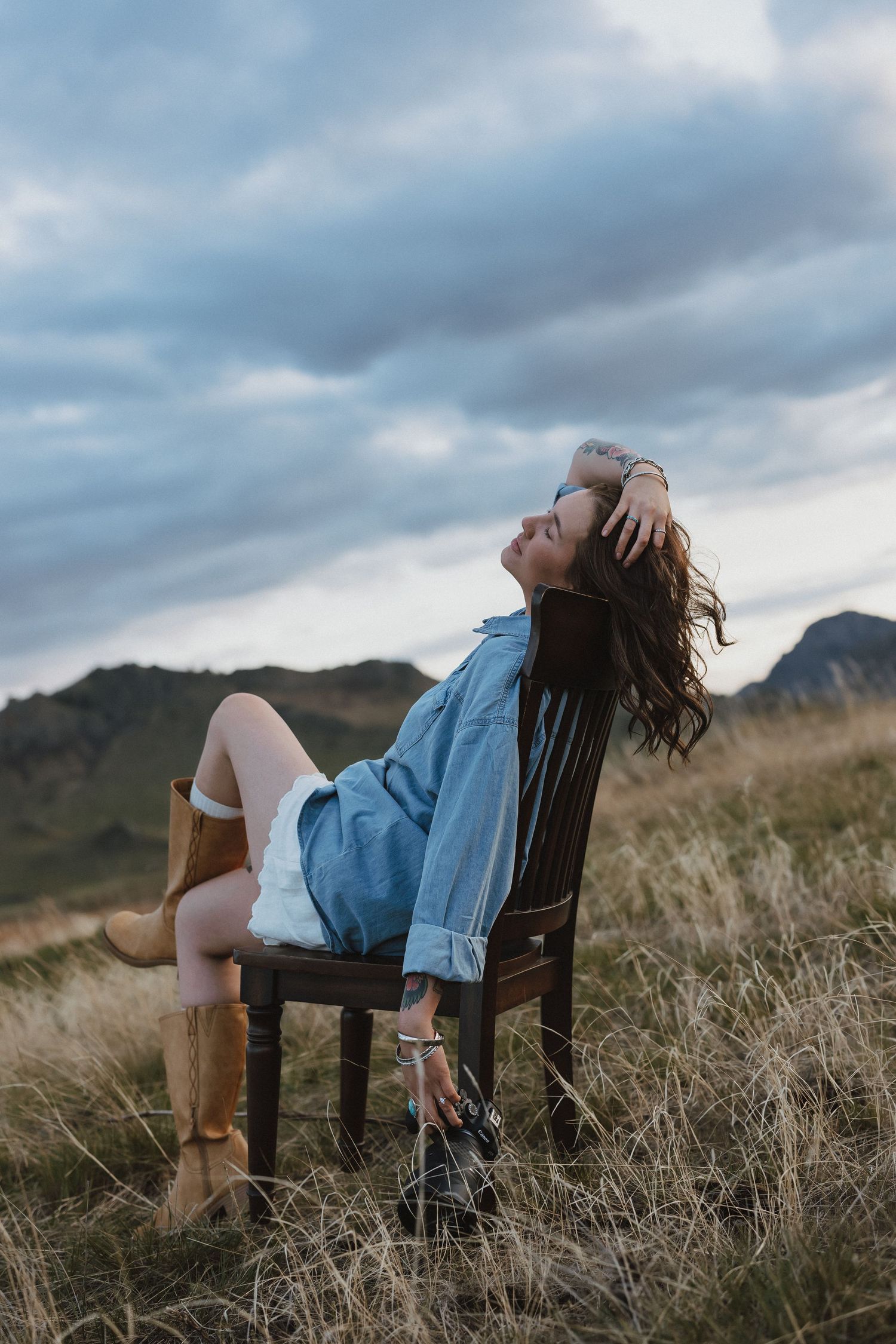 Woman in denim shirt and cowboy boots sits on wooden chair in grassy field, hair blowing in wind under cloudy sky.