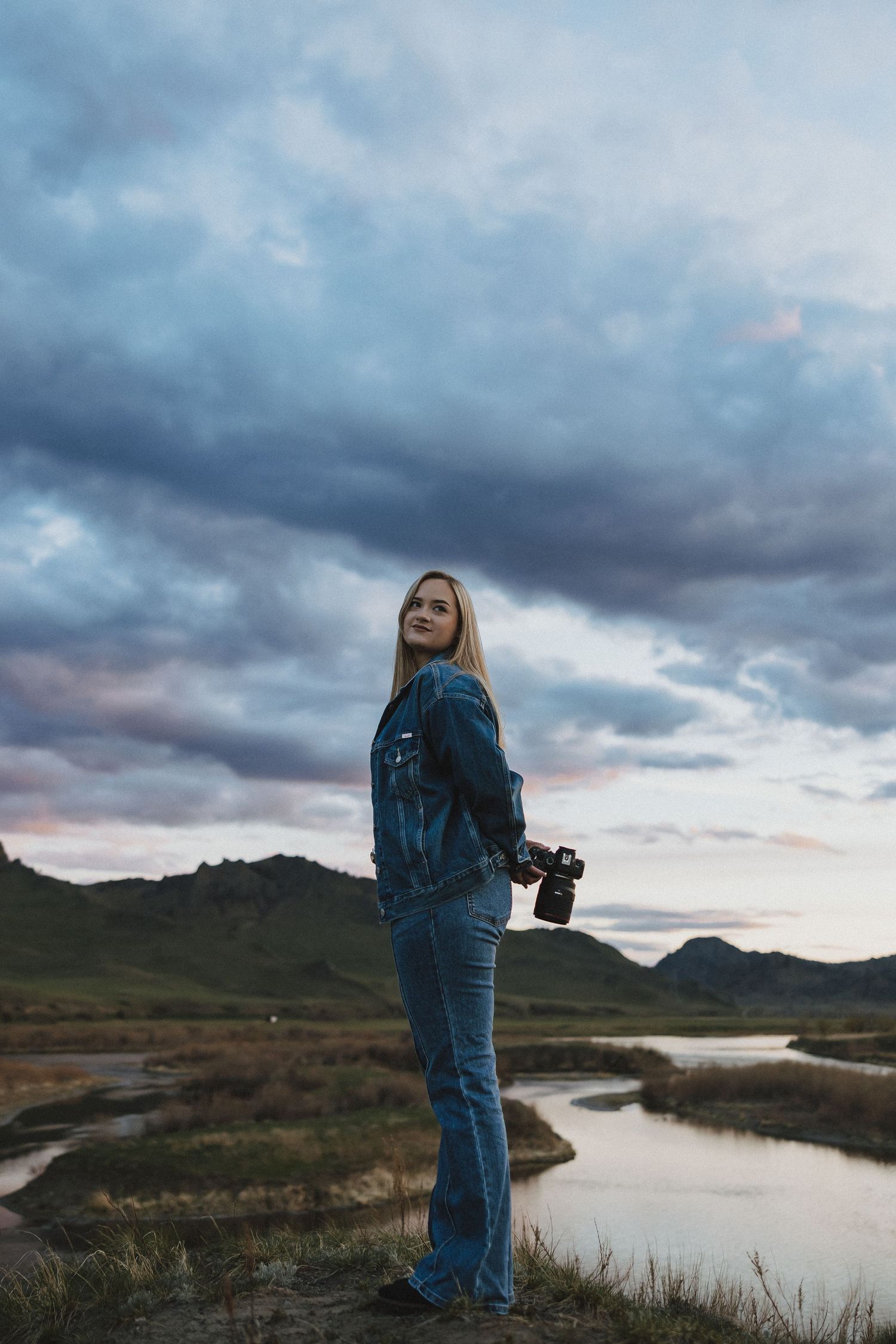 Blonde woman in denim jacket holding camera stands by winding river with dramatic cloudy sky and green hills backdrop.