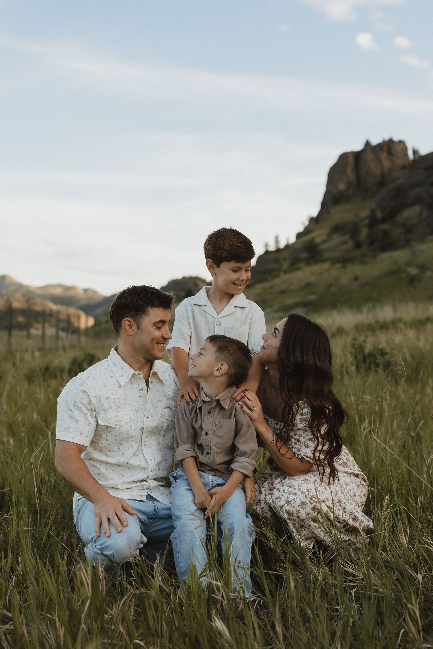 Family of four sitting in a grassy mountain meadow, smiling and laughing together on a sunny day.